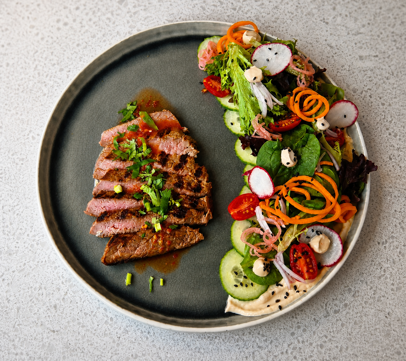 A round gray plate with sliced seared tuna topped with chopped green onions and sauce on the left side, and a colorful salad with sliced radishes, cherry tomatoes, cucumber, leafy greens, shredded carrots, and dressing on the right side.