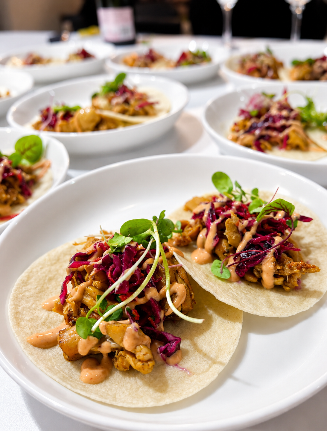 Close-up of two tacos filled with seasoned meat, red cabbage slaw, and microgreens on a white plate at a banquet or restaurant.