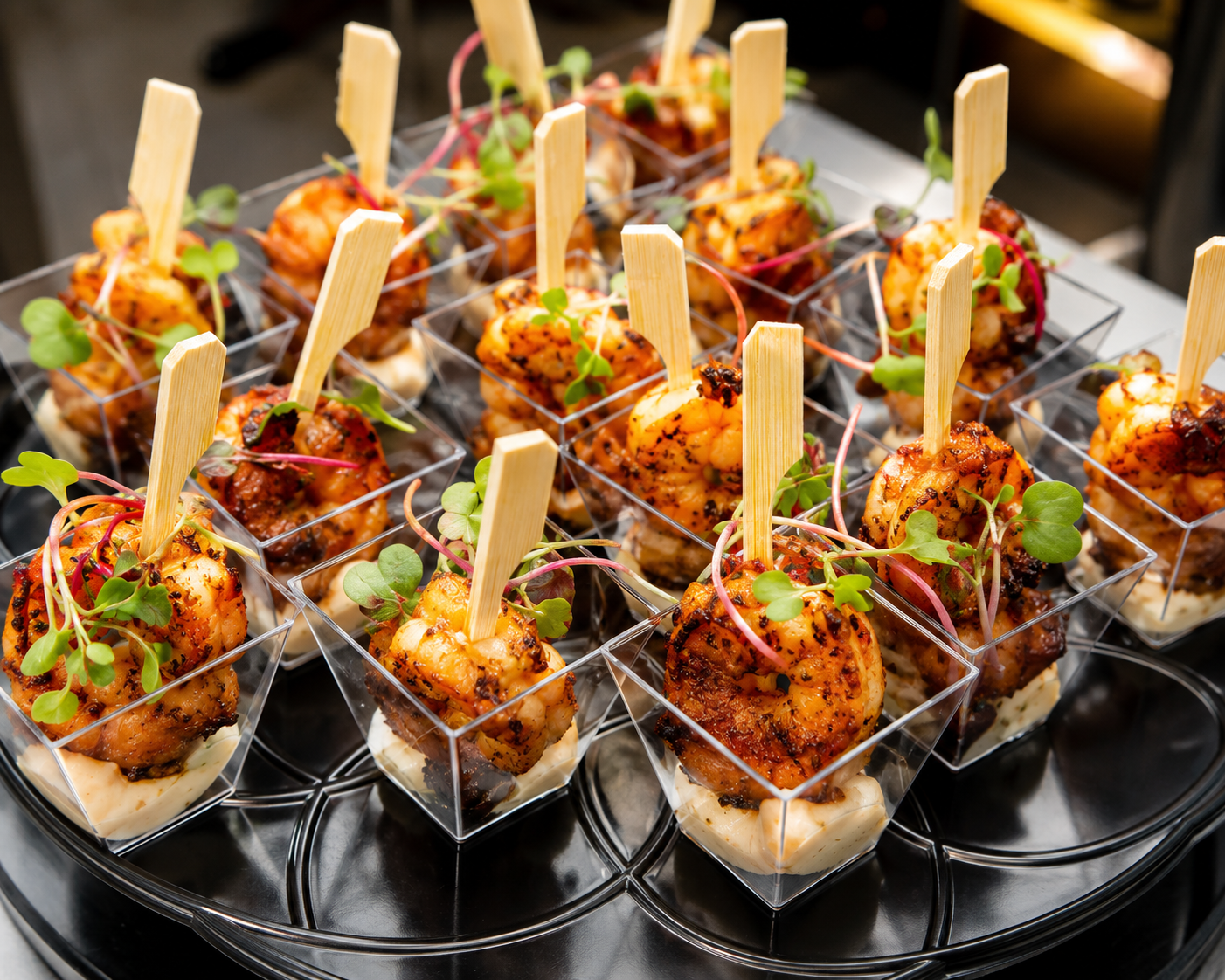 Appetizers of grilled shrimp skewers garnished with microgreens served in clear plastic cups on a black tray.
