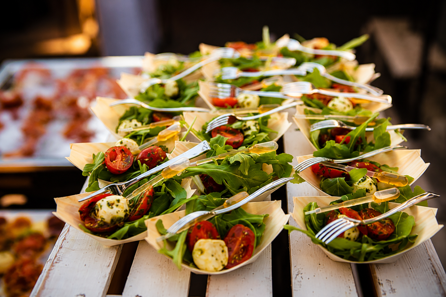 Assorted fresh salads with cherry tomatoes, mozzarella balls, and greens served in small wooden boats with forks, set on a white wooden surface.