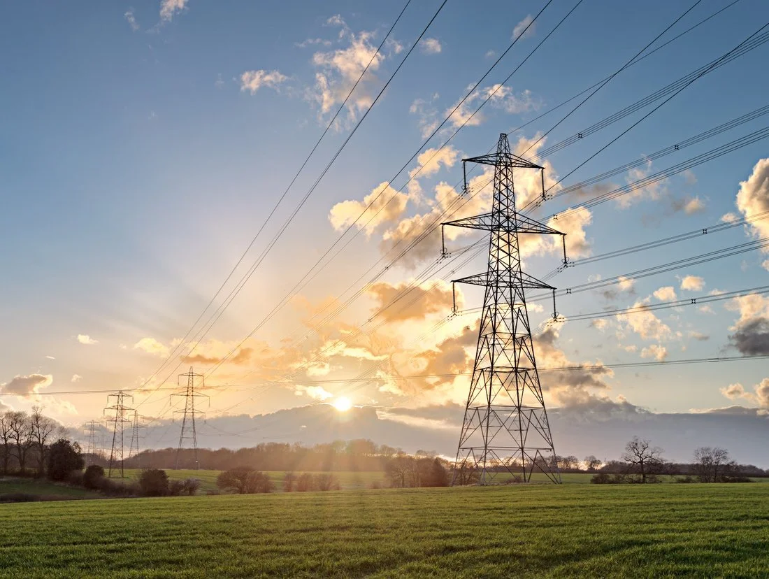 Ssun setting behind high-voltage power lines and transmission towers in a rural area with green fields and trees.