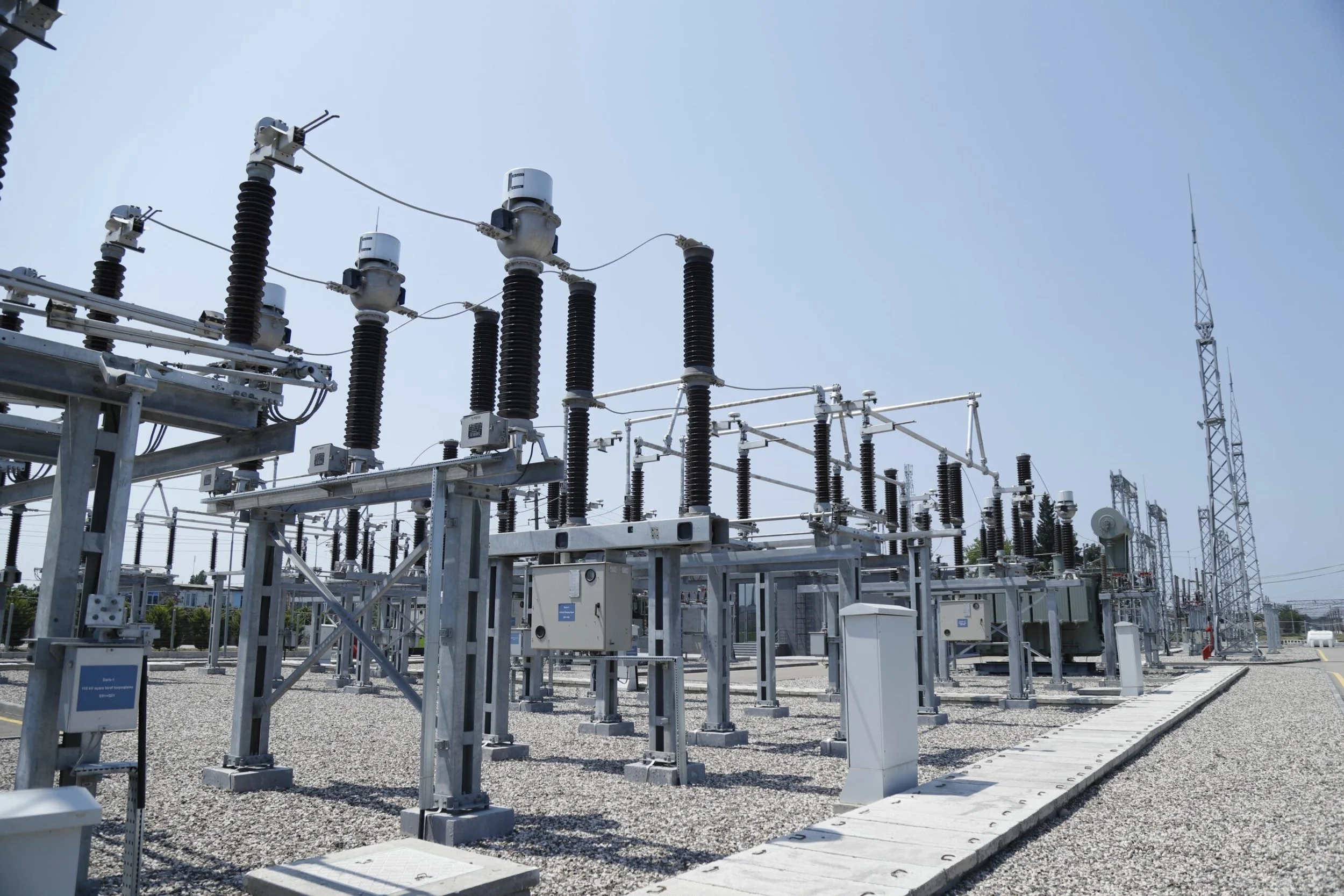 An electrical substation with power transformers, insulators, and transmission towers under a clear sky.