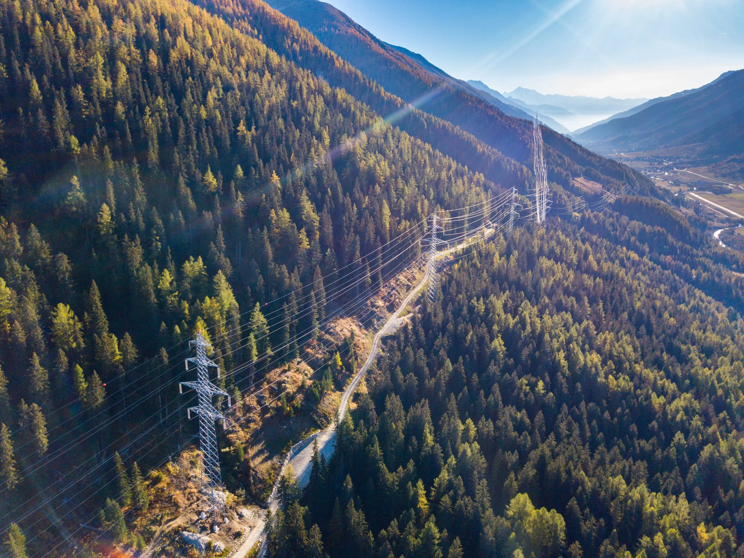 Aerial view of a mountainous landscape with a dense forest of green trees, a winding dirt road, and power lines running through the valley. The sky is clear and sunny.
