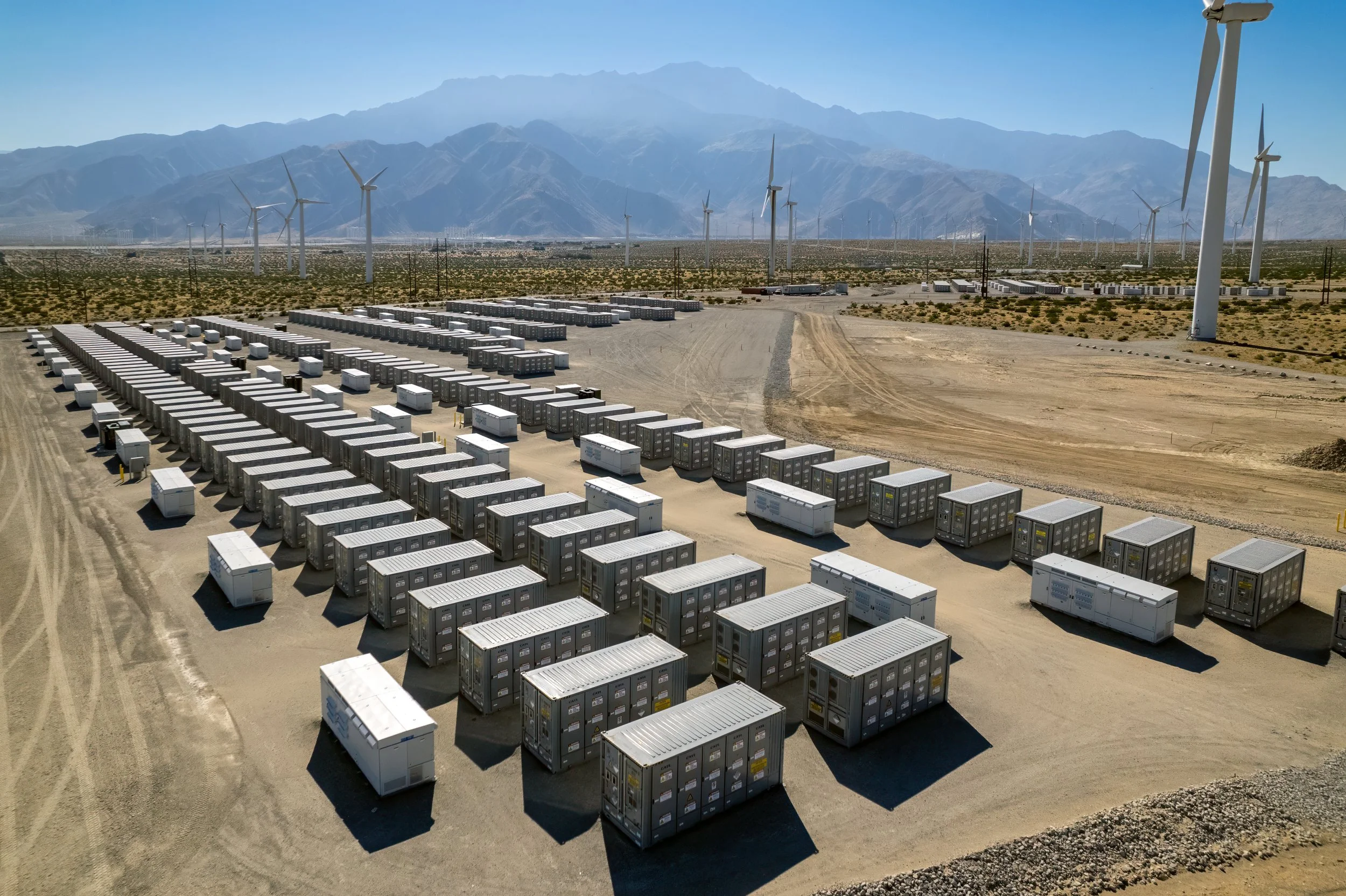 A large renewable energy storage facility with numerous shipping containers, wind turbines, and mountains in the background.