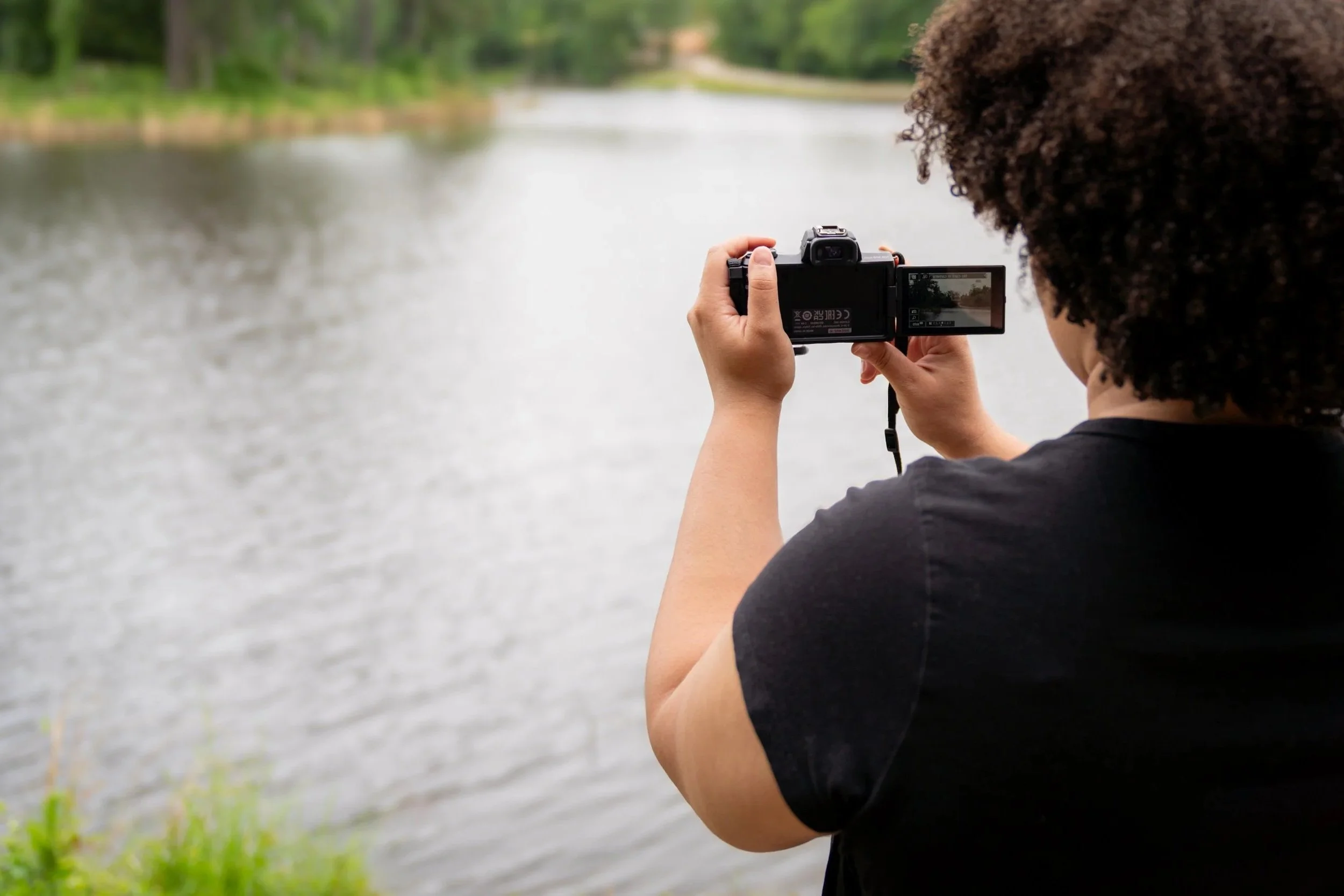 Person with curly hair taking a photo with a camera by a river or lake, surrounded by greenery.
