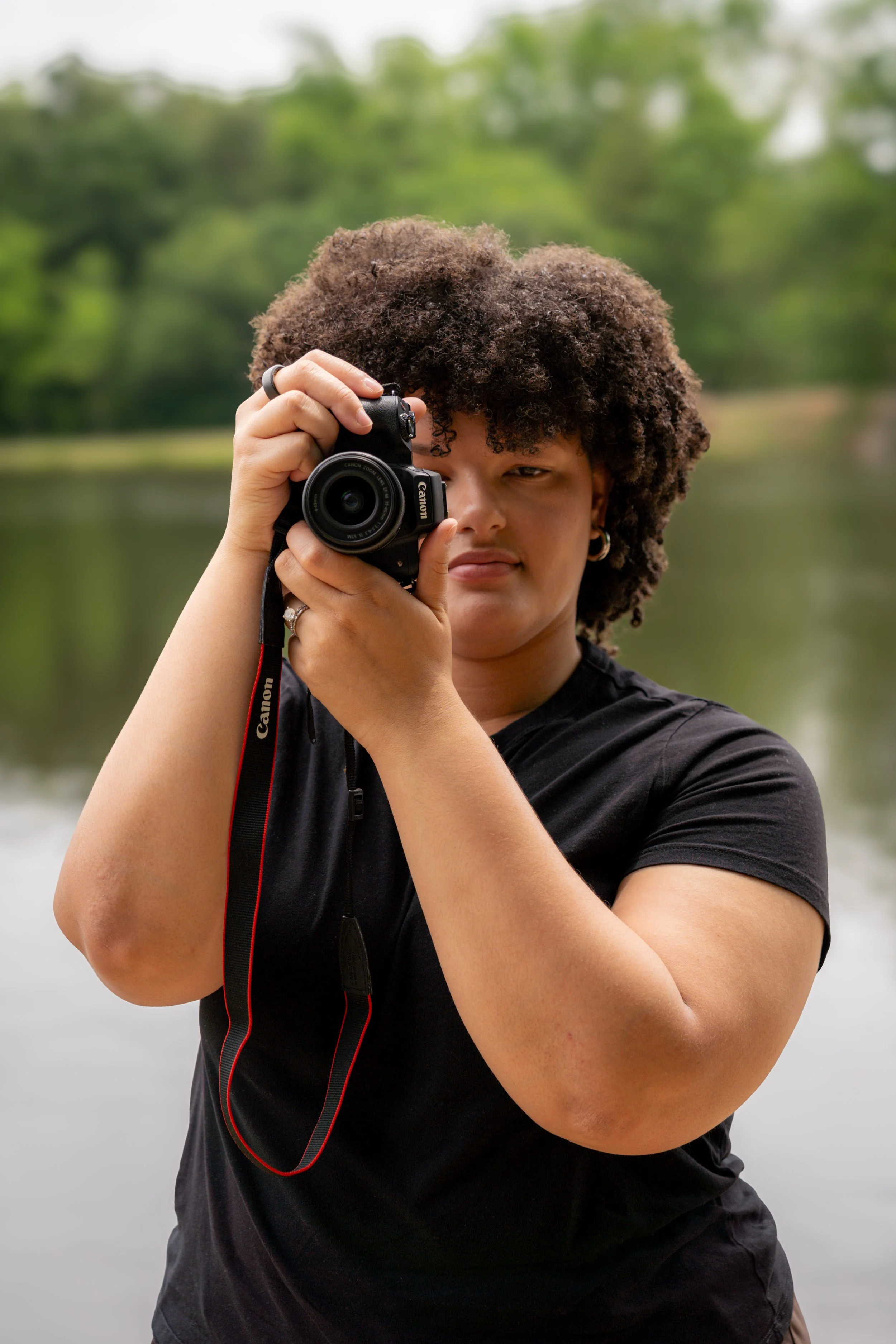 A woman with curly hair holding a camera close to her face, outdoors with blurred green trees and water in the background.