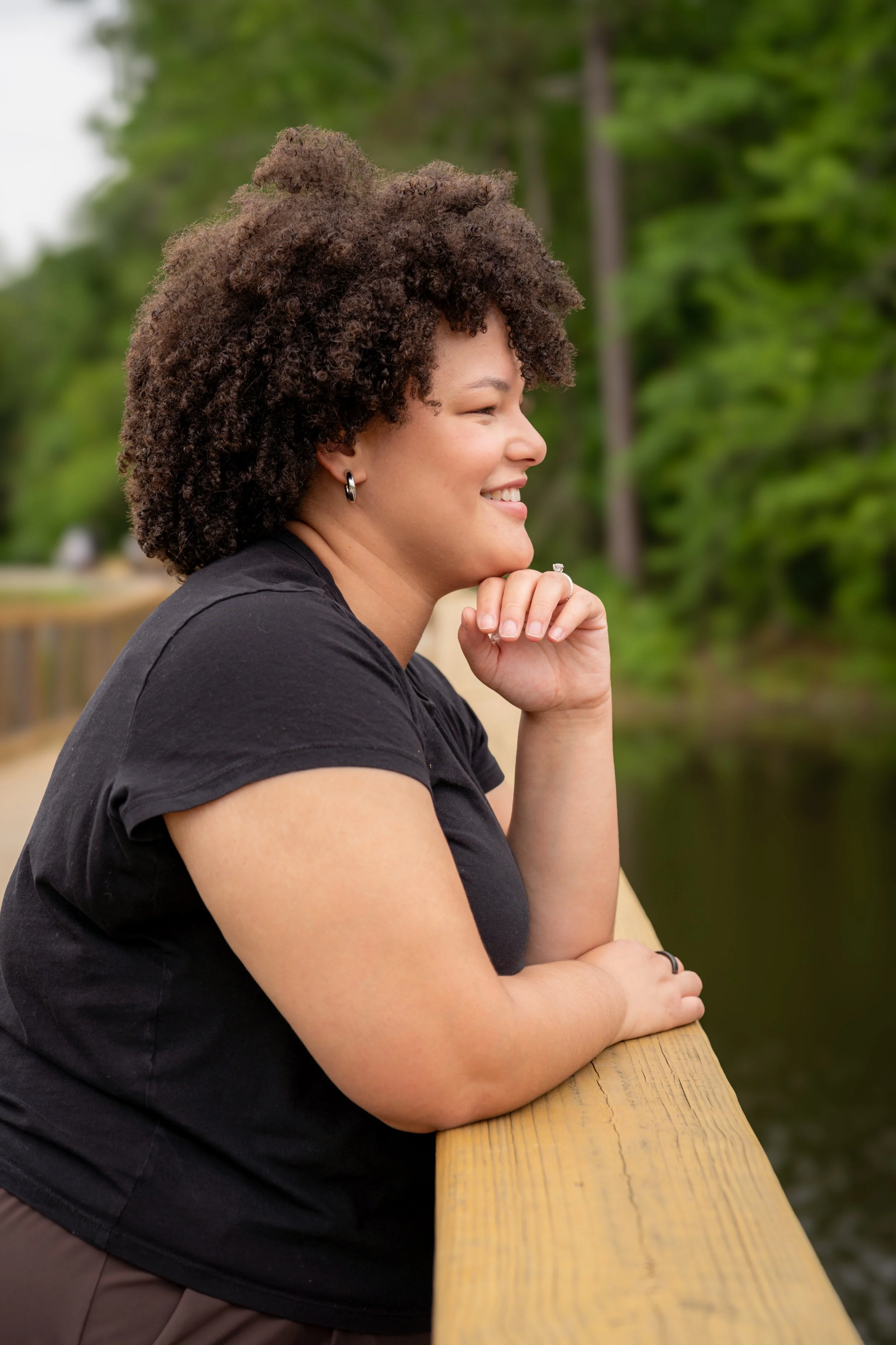 Side view of a woman with dark brown curly hair smiling softly, resting her chin on her hand, leaning on a wooden railing outdoors with greenery in the background.