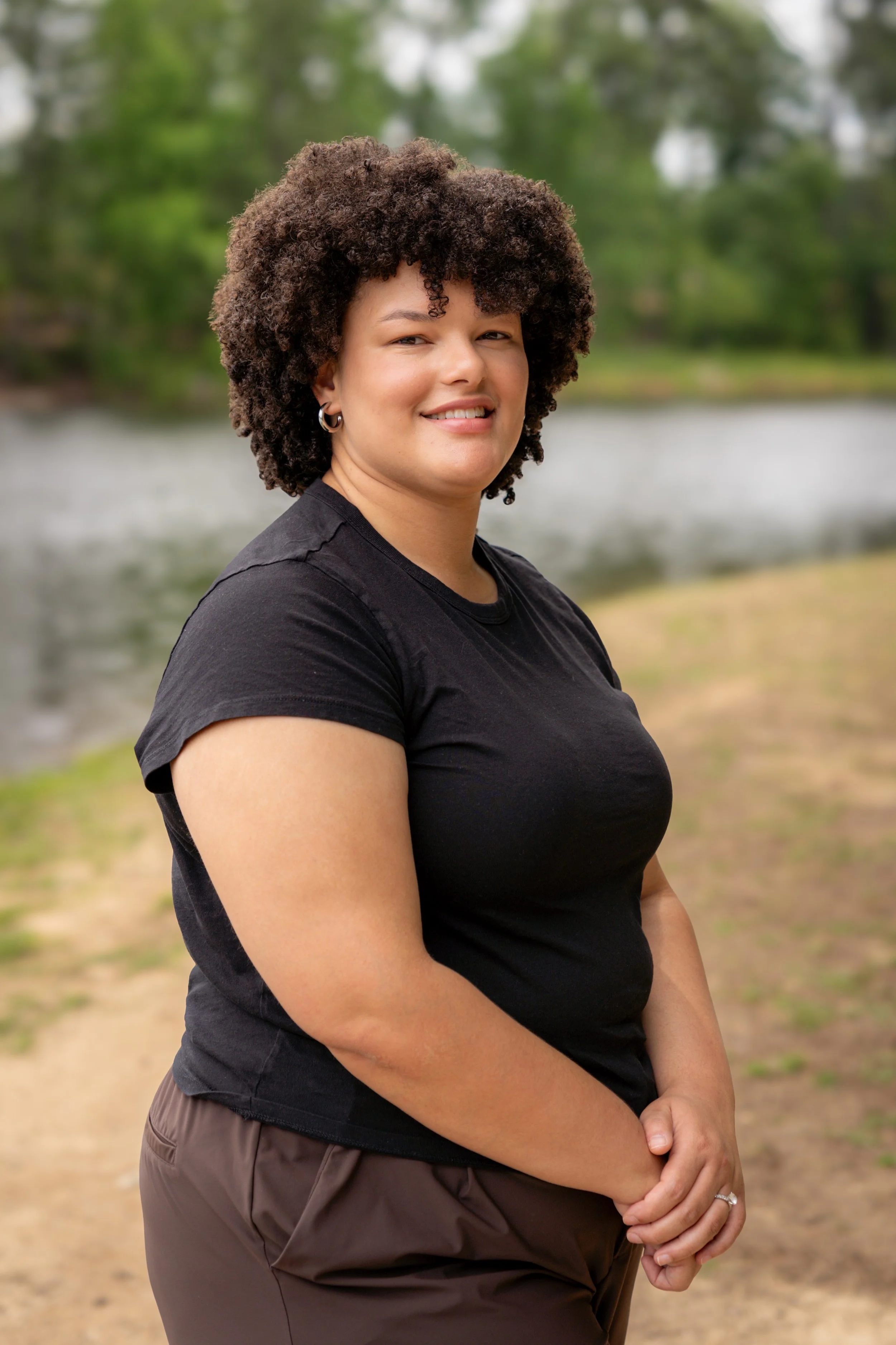 A woman with curly hair smiling outdoors near a body of water with trees in the background.