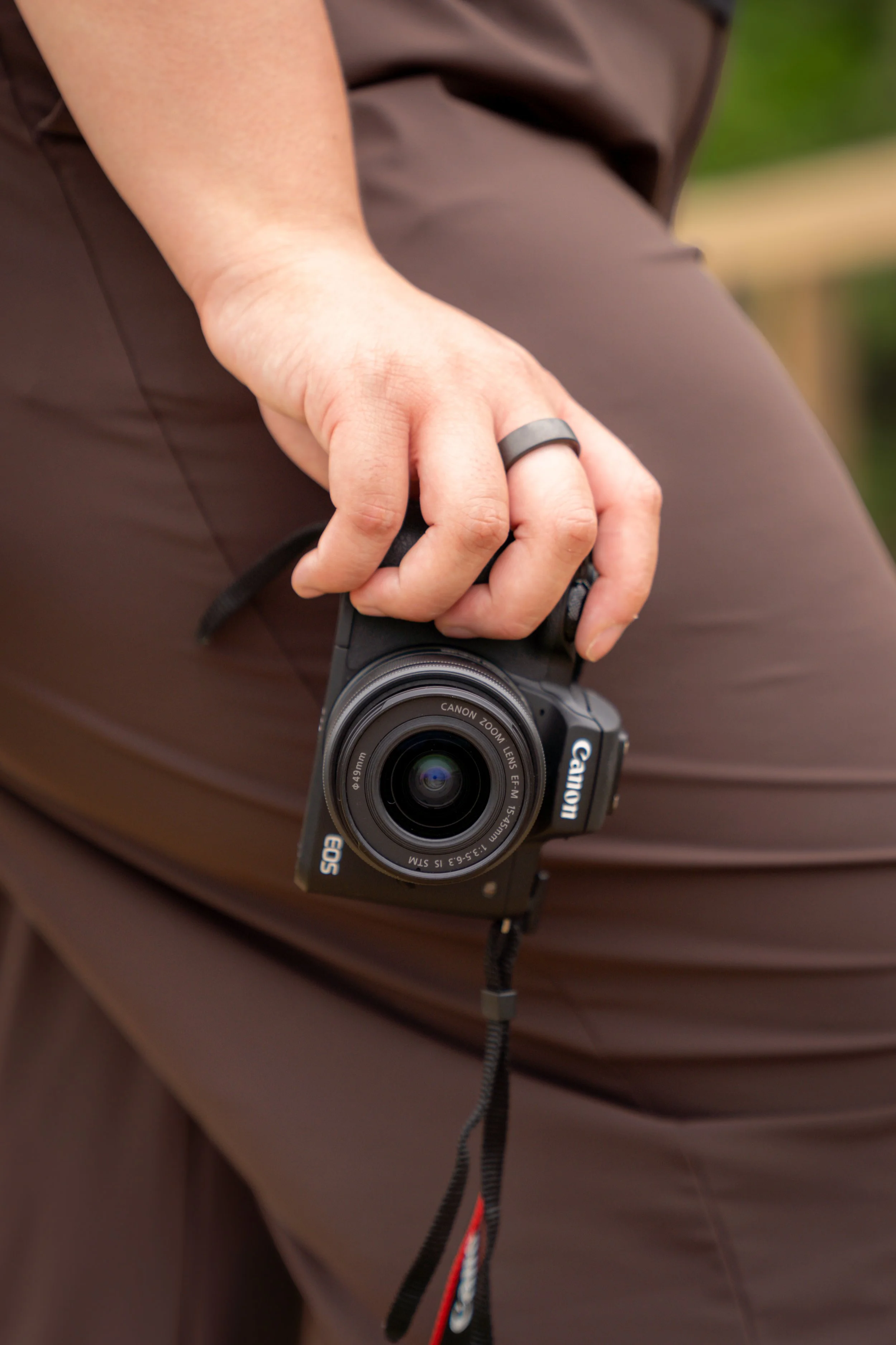 Close-up of a person's hand holding a Canon point-and-shoot camera, wearing a black ring on the middle finger and a ring on the pinky finger, with a brown garment in the background.