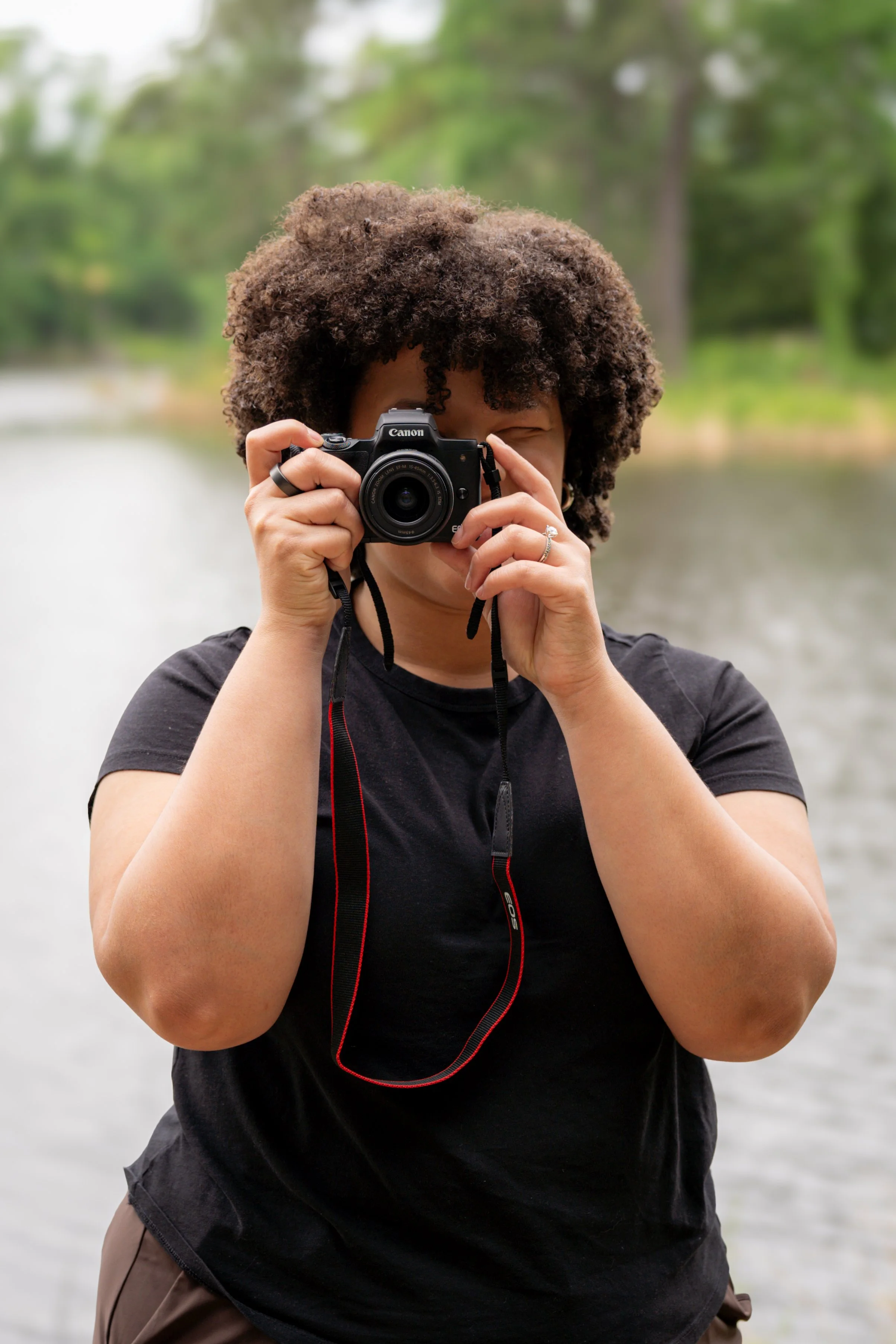 A person with curly hair taking a photograph outdoors near a body of water with trees in the background.