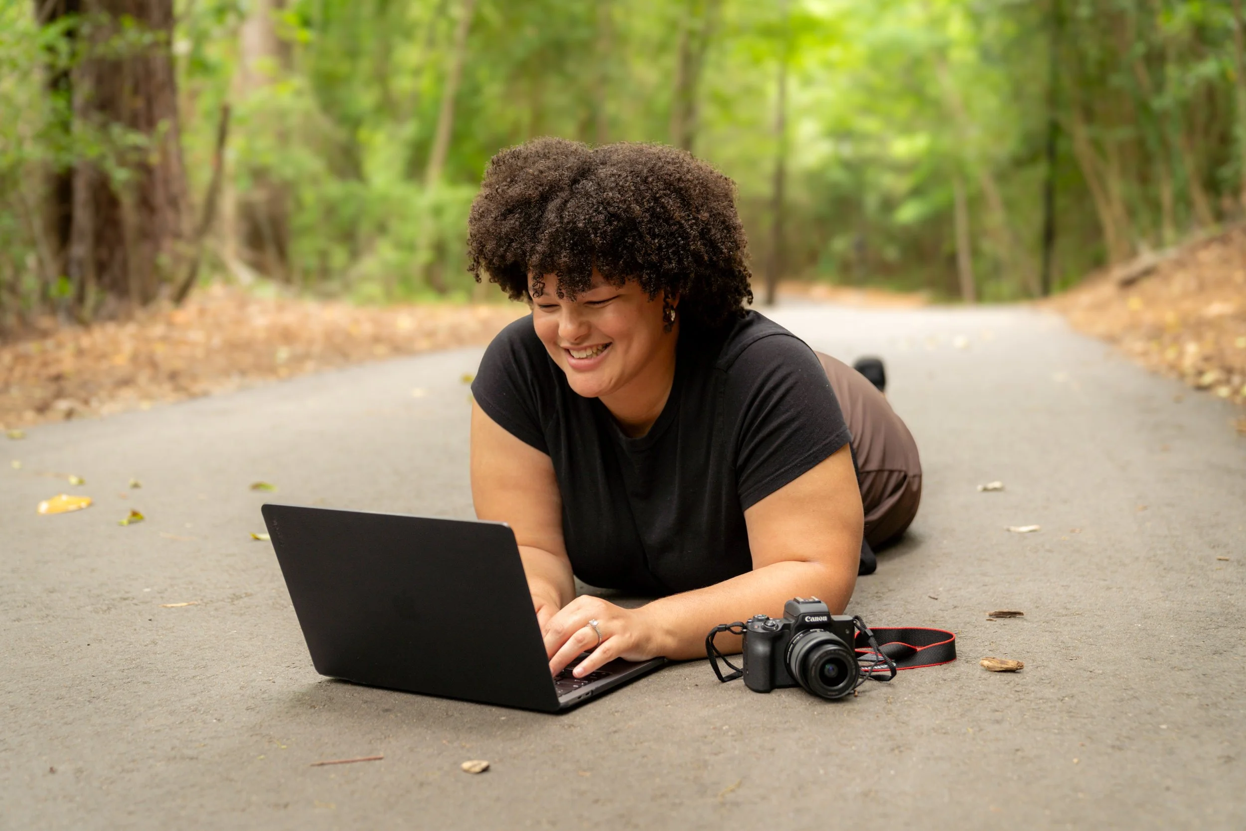 Woman with curly hair smiling while lying on her stomach on a paved forest path, using a laptop, with camera on the ground beside her.