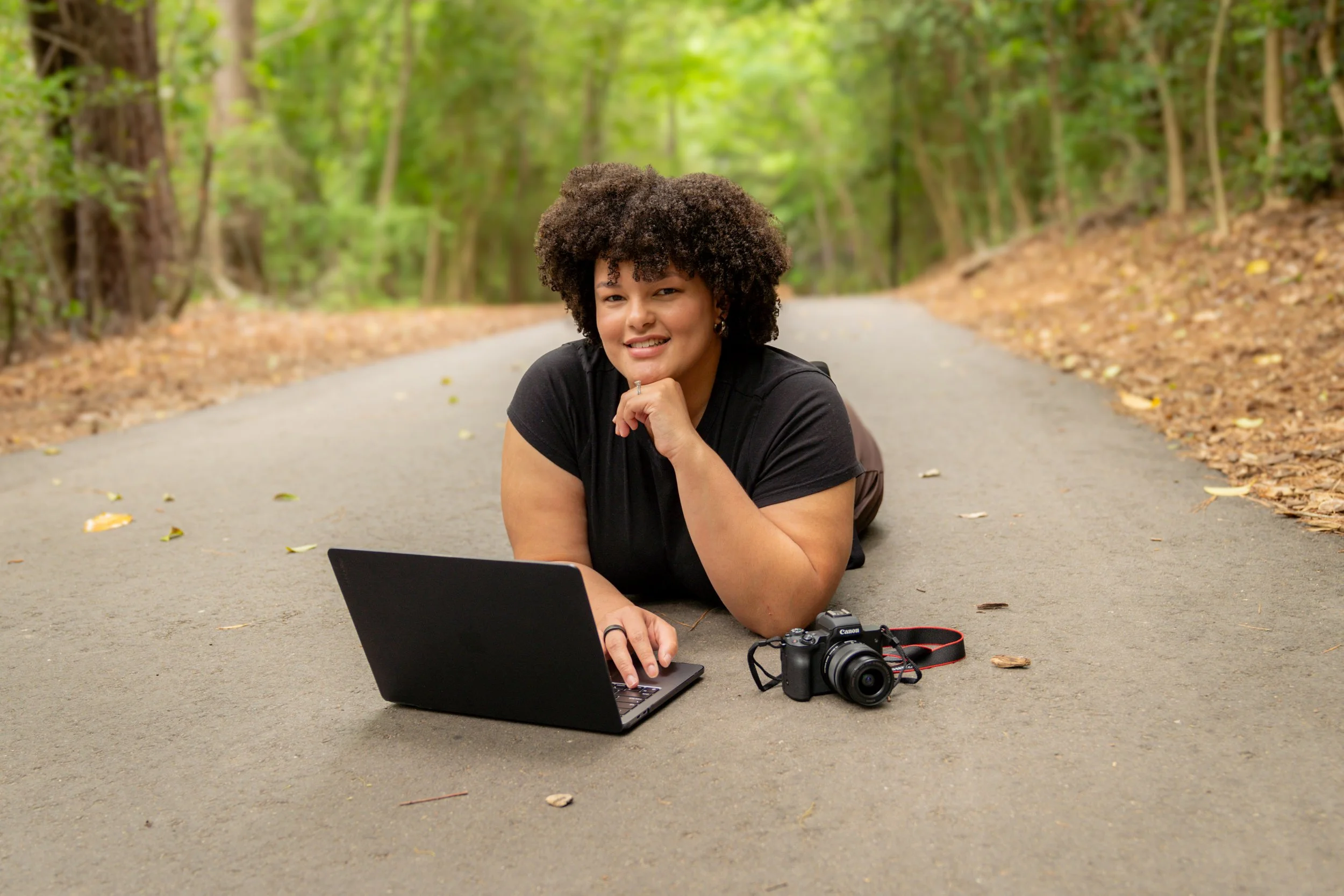 A woman with curly hair lying on the road in a forest, working on a laptop, with a camera beside her.