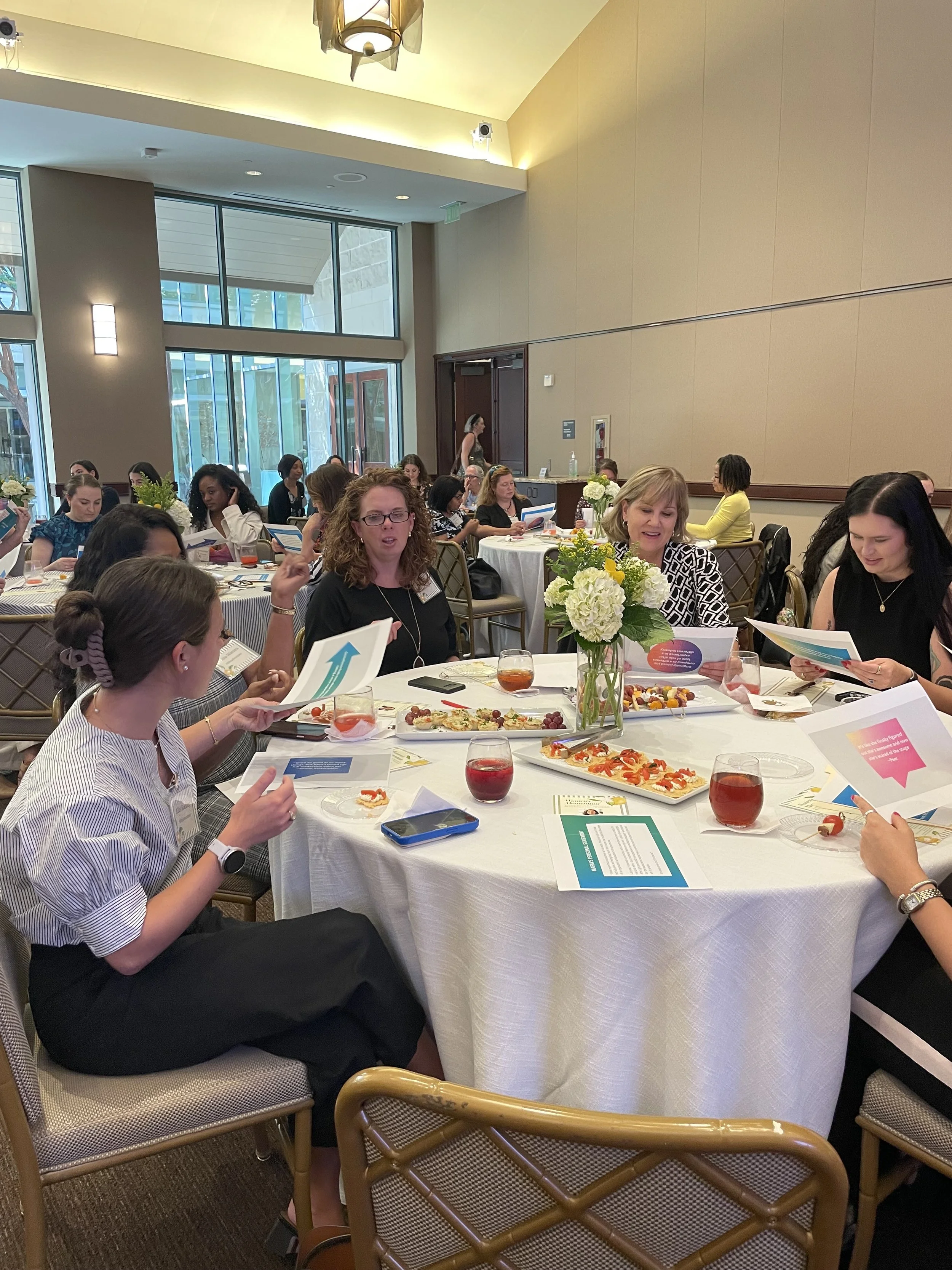 This AMPlify Coaching and Consulting workshop shows a group of women engaging in conversation at a table. The group is one of many groups in the room, and all groups are discussing strategies for improving professional branding.