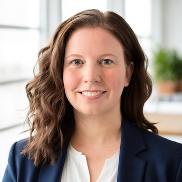 Allison Pickett is an executive coach and strategy consultant. In this picture, she is has shoulder-length red hair , is wearing a navy blazer and white blouse, and is smiling in a bright office setting.