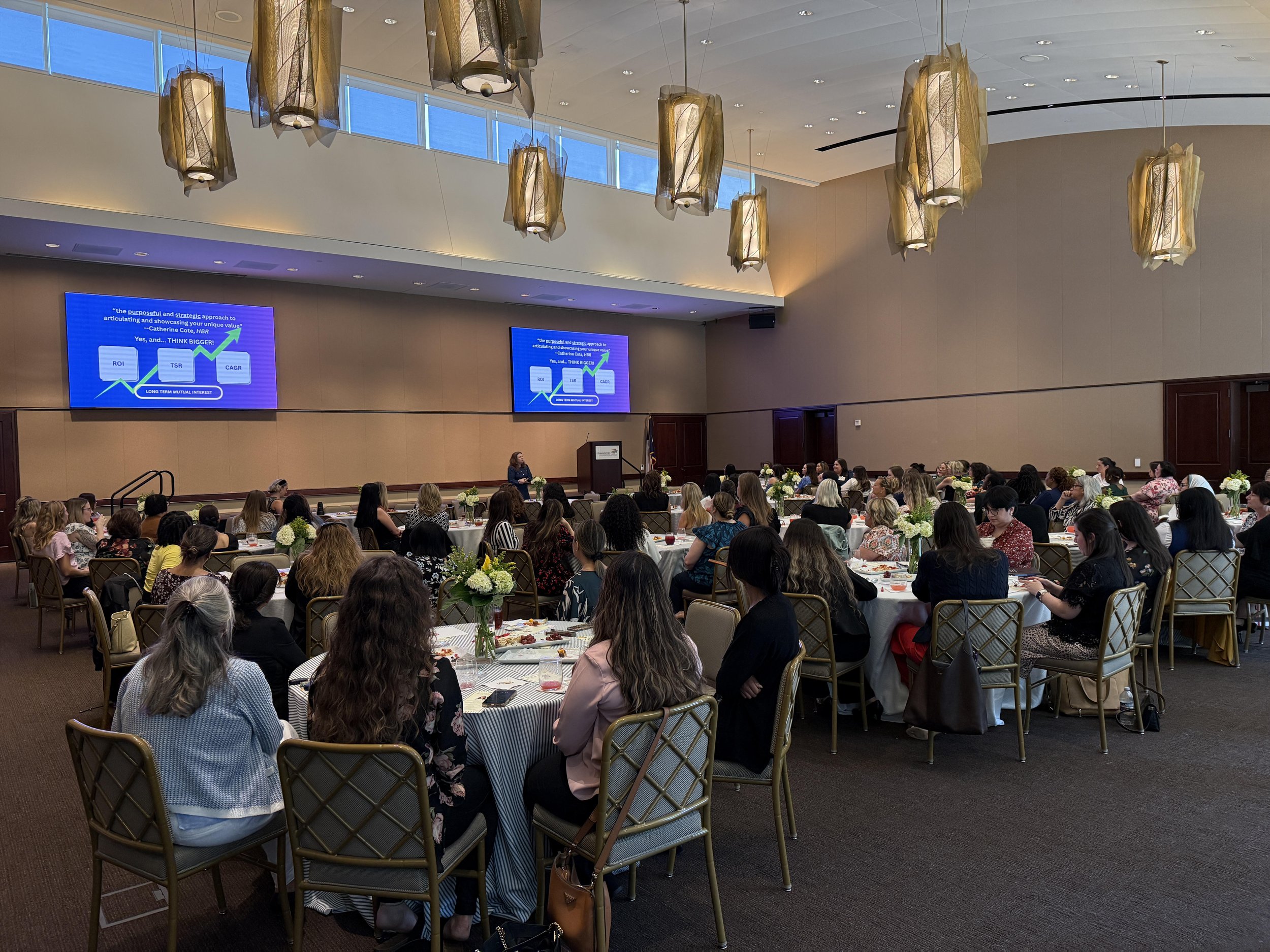 Executive coach Allison Pickett is speaking to a group of about 150 women at a professional branding conference. Groups of 8-10 women are at each table. A presentation slide is projected on two screens displaying a chart related to business metrics.