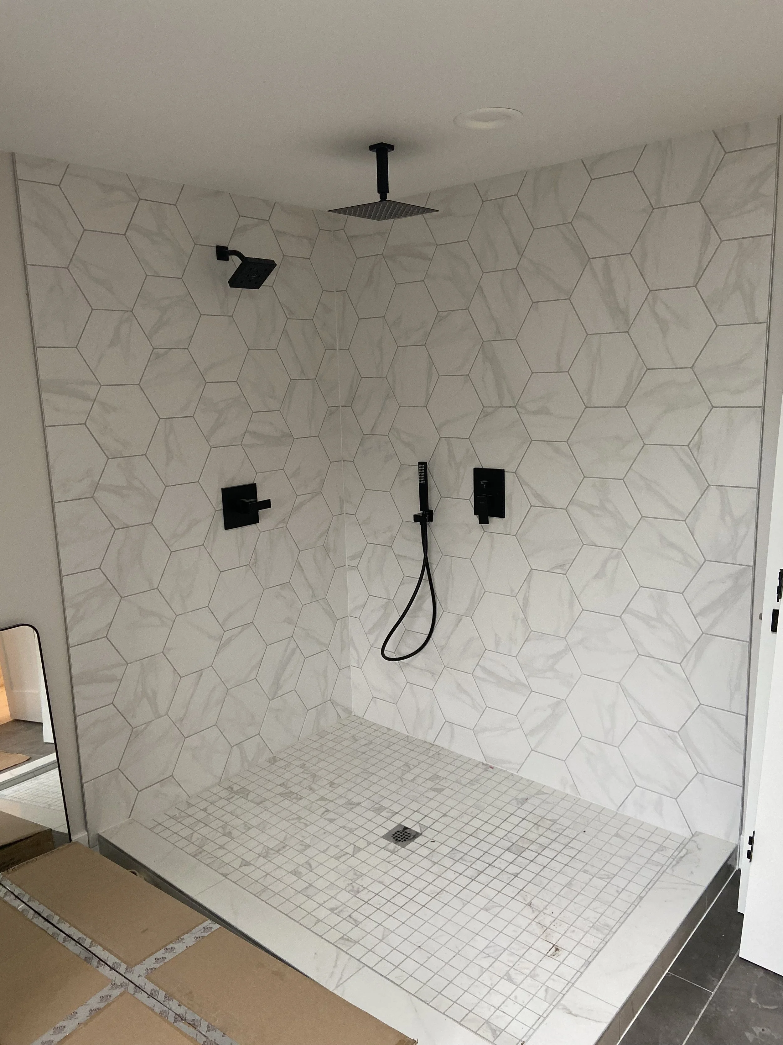 Bathroom shower with white marble hexagonal wall tiles, a rain showerhead, a wall-mounted handheld shower, and a drain in the tiled shower floor.