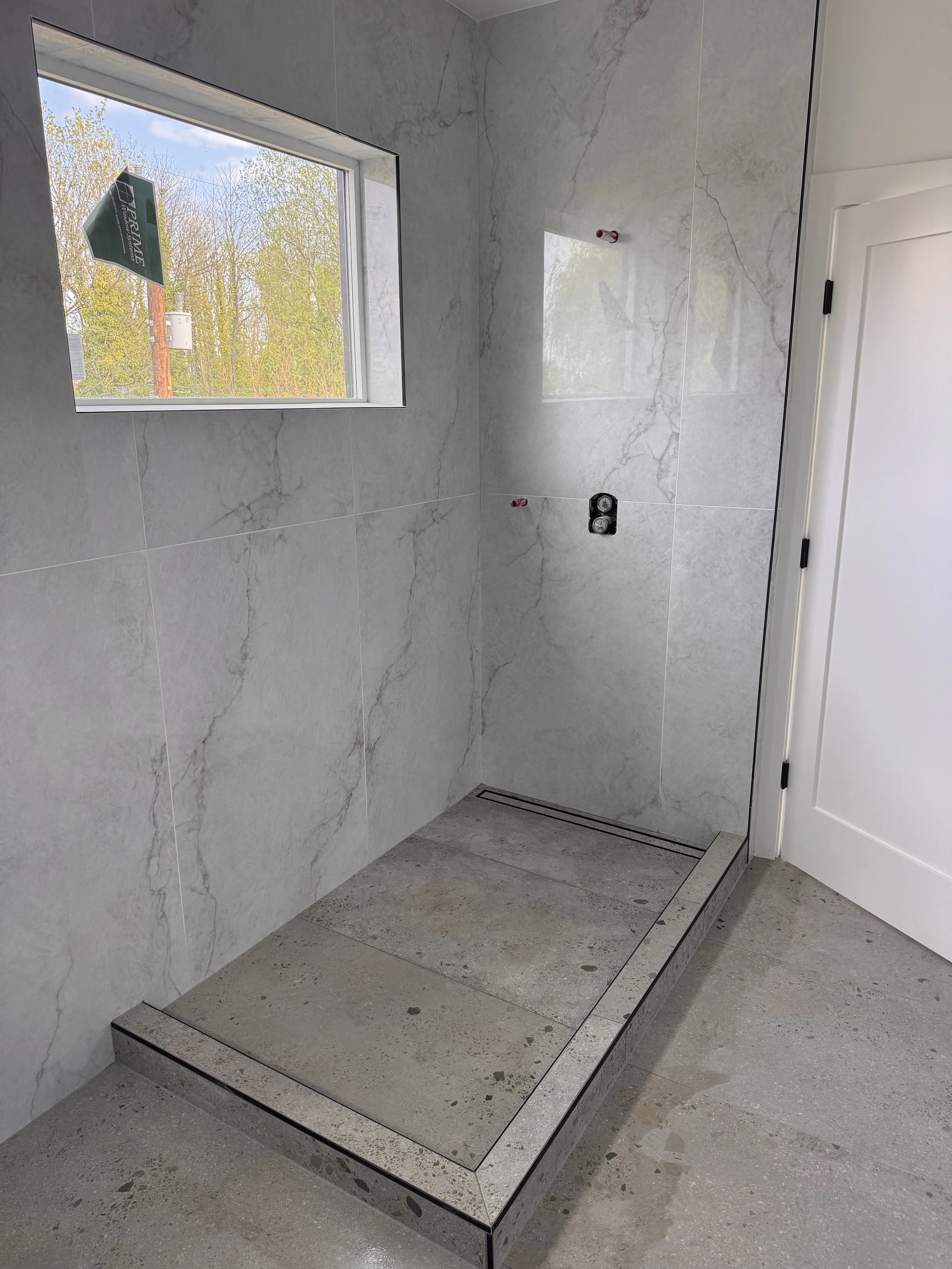 An empty walk-in shower with light gray marble tiles, a small window overlooking trees, a drainage grate on the floor, and a door on the right side.
