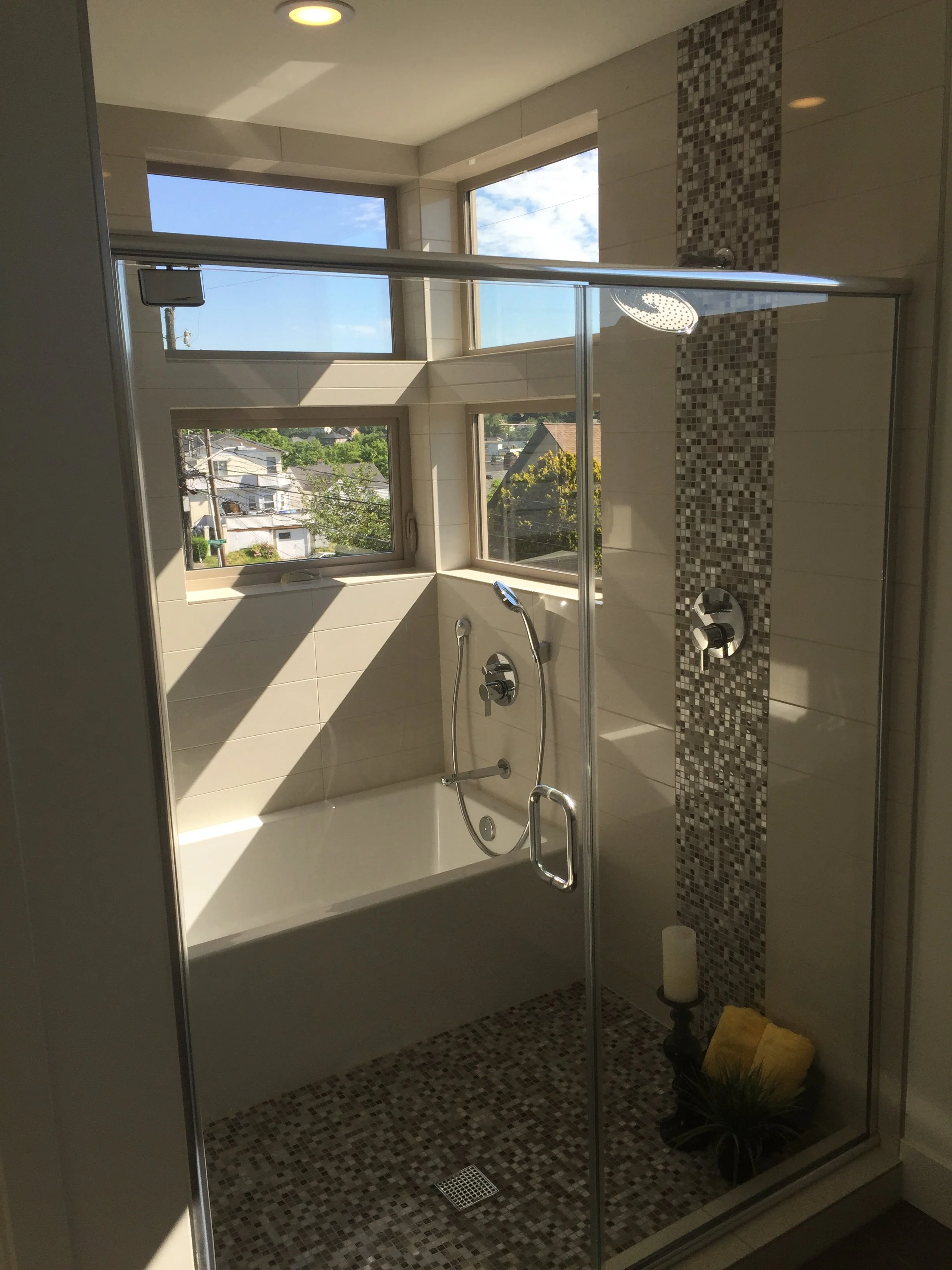 Modern walk-in shower with glass enclosure, a rainfall showerhead, a window showing a neighborhood outside, black and beige mosaic tiles, and a yellow towel and decorative plant on the floor.