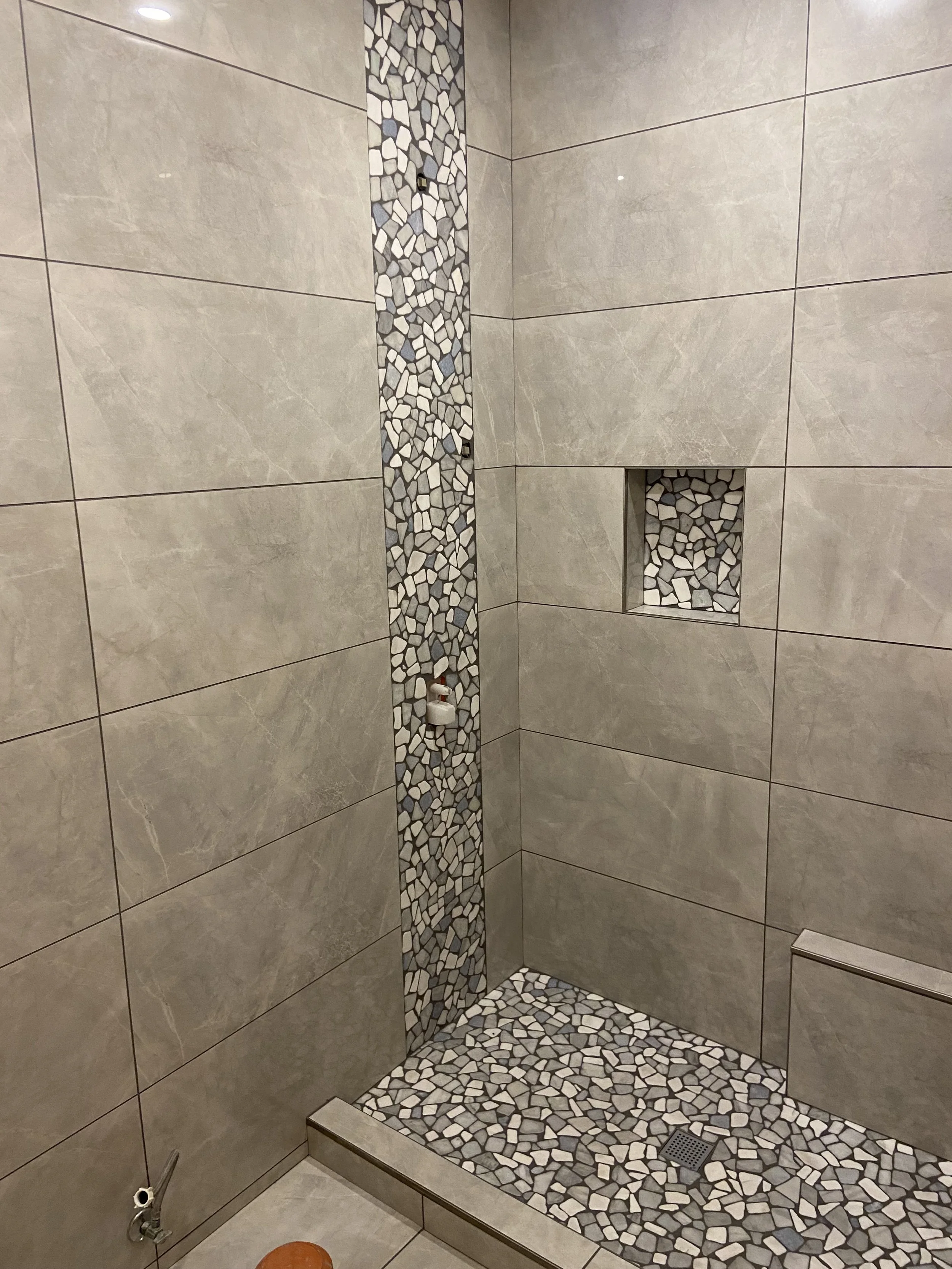 A corner of a shower with large beige tiles on the walls, a vertical strip of small pebble-like tiles, a built-in niche with pebble tiles, a floor drain with pebble tiles surrounding it, and a small metal water tap on the lower left side.
