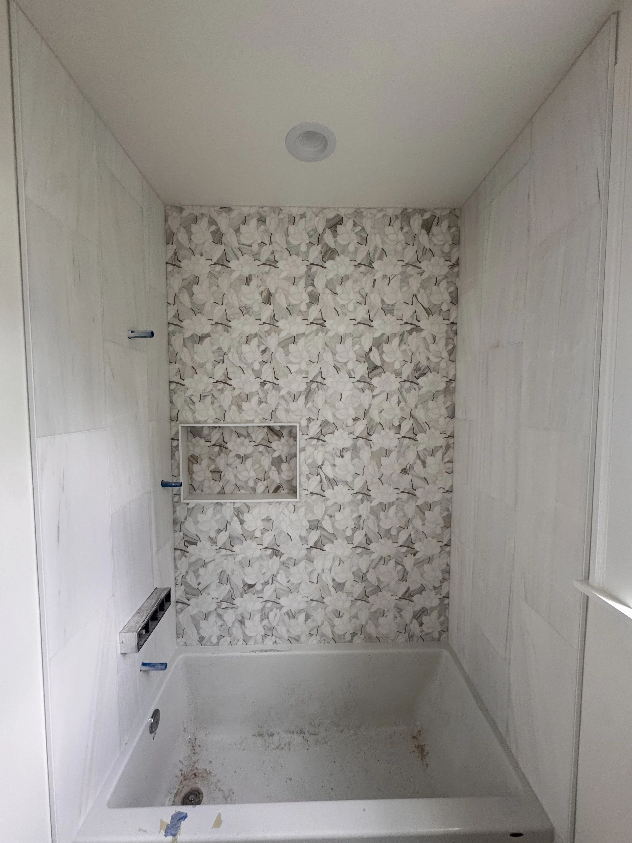 Empty shower with floral patterned tiles on the back wall, light-colored side walls, a drain, and an unfinished base with some dirt.