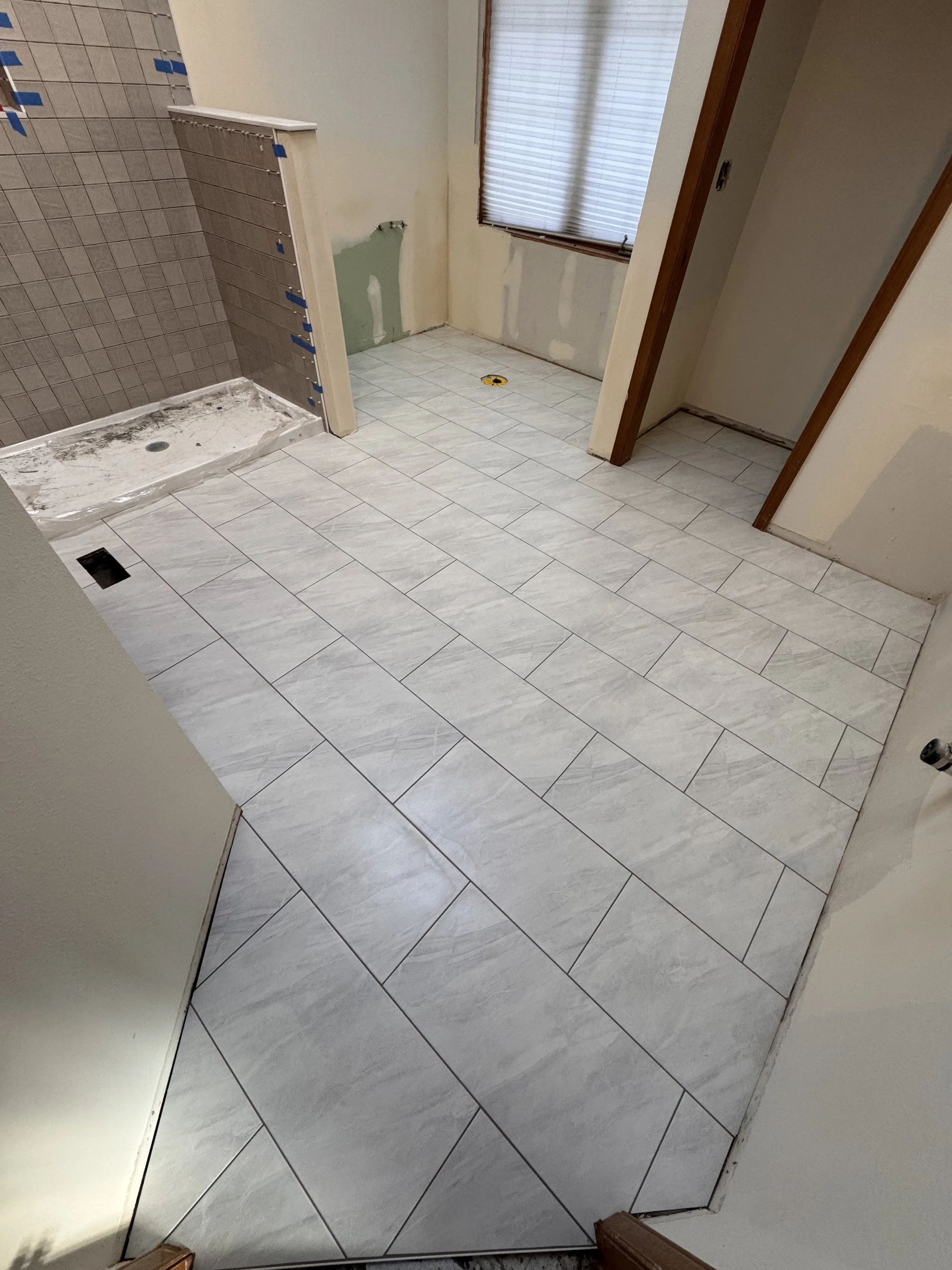 Room under renovation, with new light gray tiles on the floor, an unfinished wall with green drywall patch, a shower area with gray tiles, and a window with closed blinds.