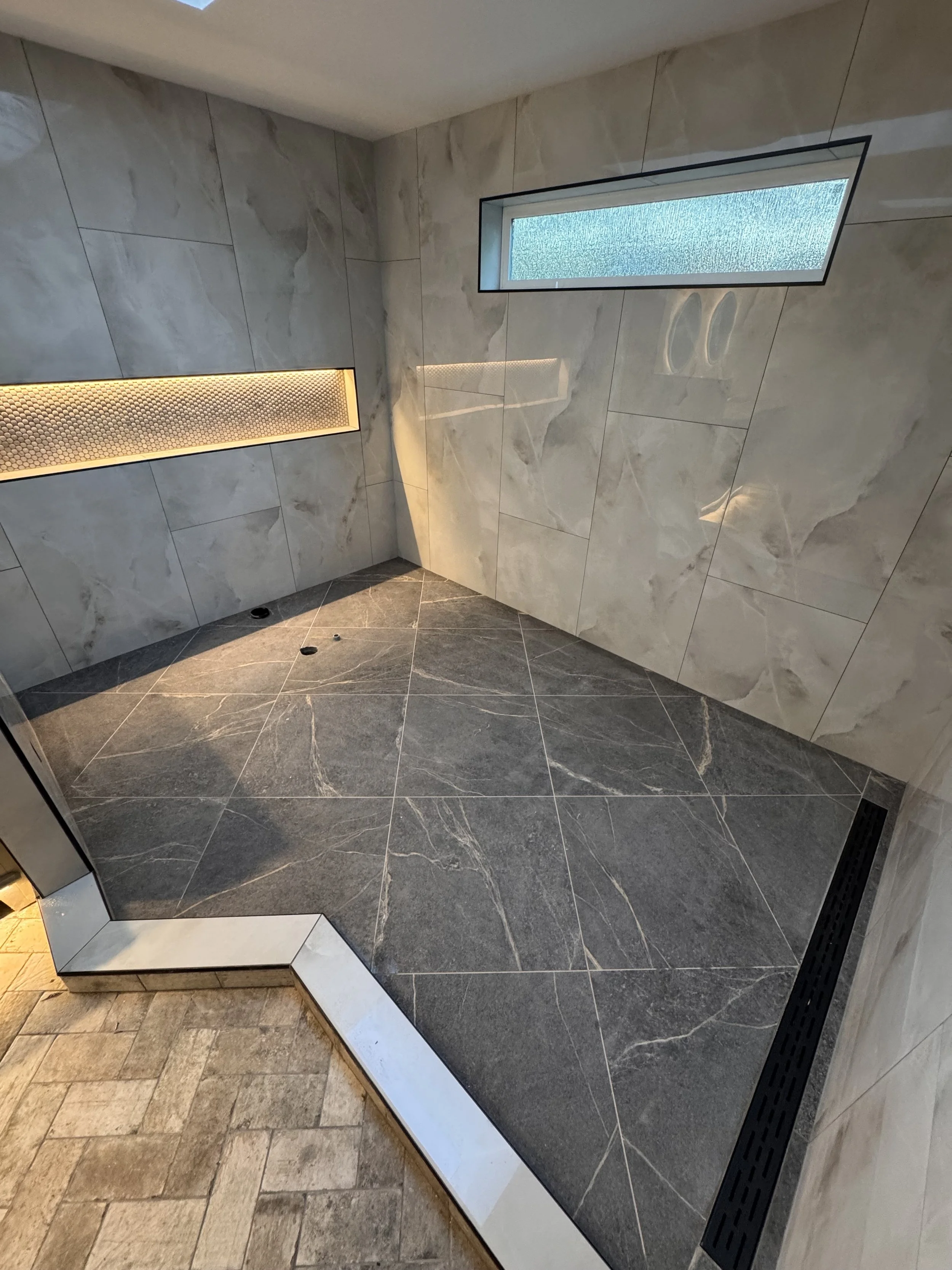 A modern, minimalist shower area with large gray marble tiles on the walls and a tankless shower drain. There is a rectangular frosted window on the right wall and a hidden linear LED light on the left wall providing warm illumination.