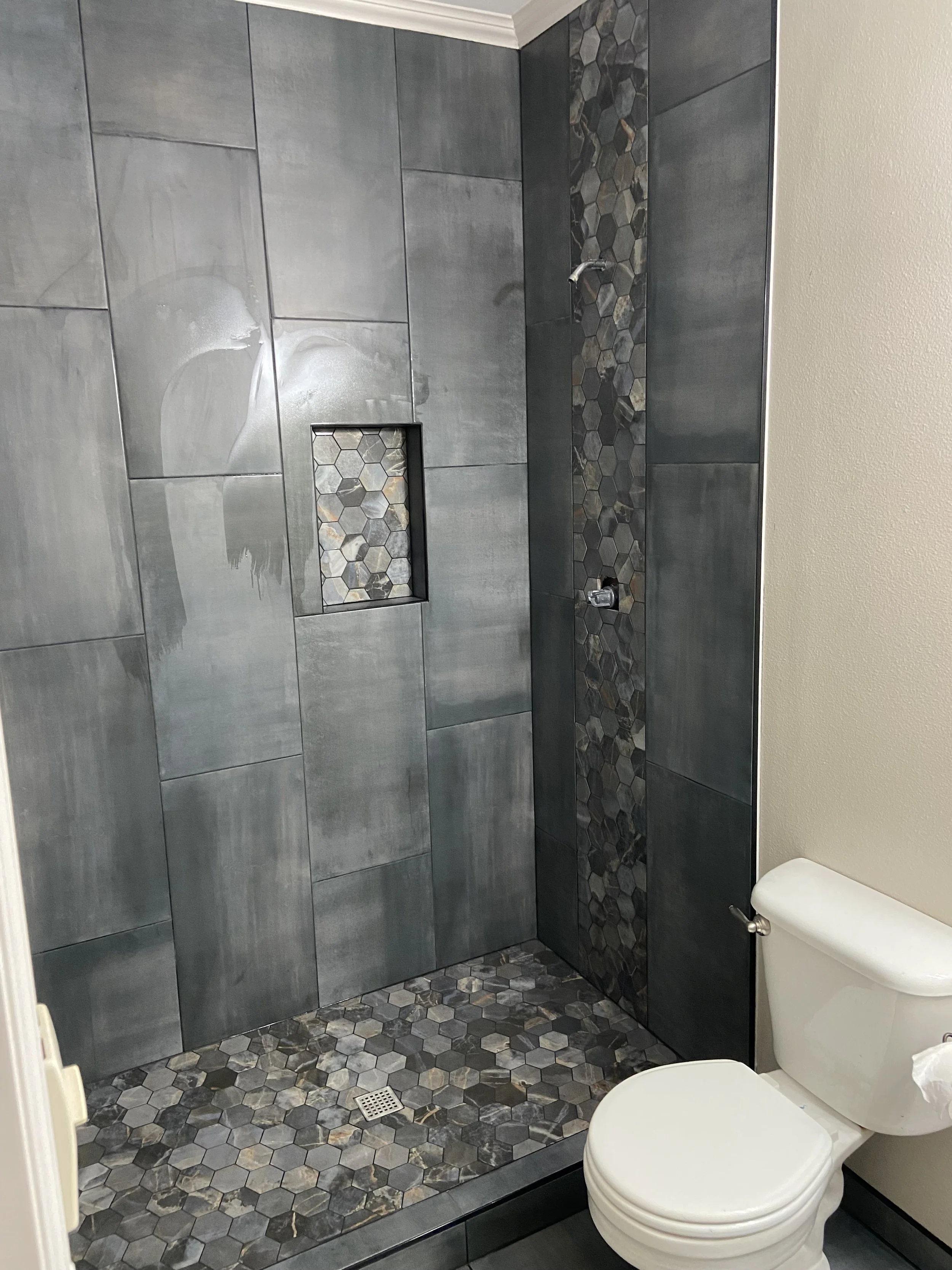 Bathroom with a walk-in shower featuring gray tile walls and a decorative strip of multicolored hexagonal tiles. The shower floor has matching hexagonal tiles, and there is a small built-in shelf with the same tile pattern. A toilet is visible in the