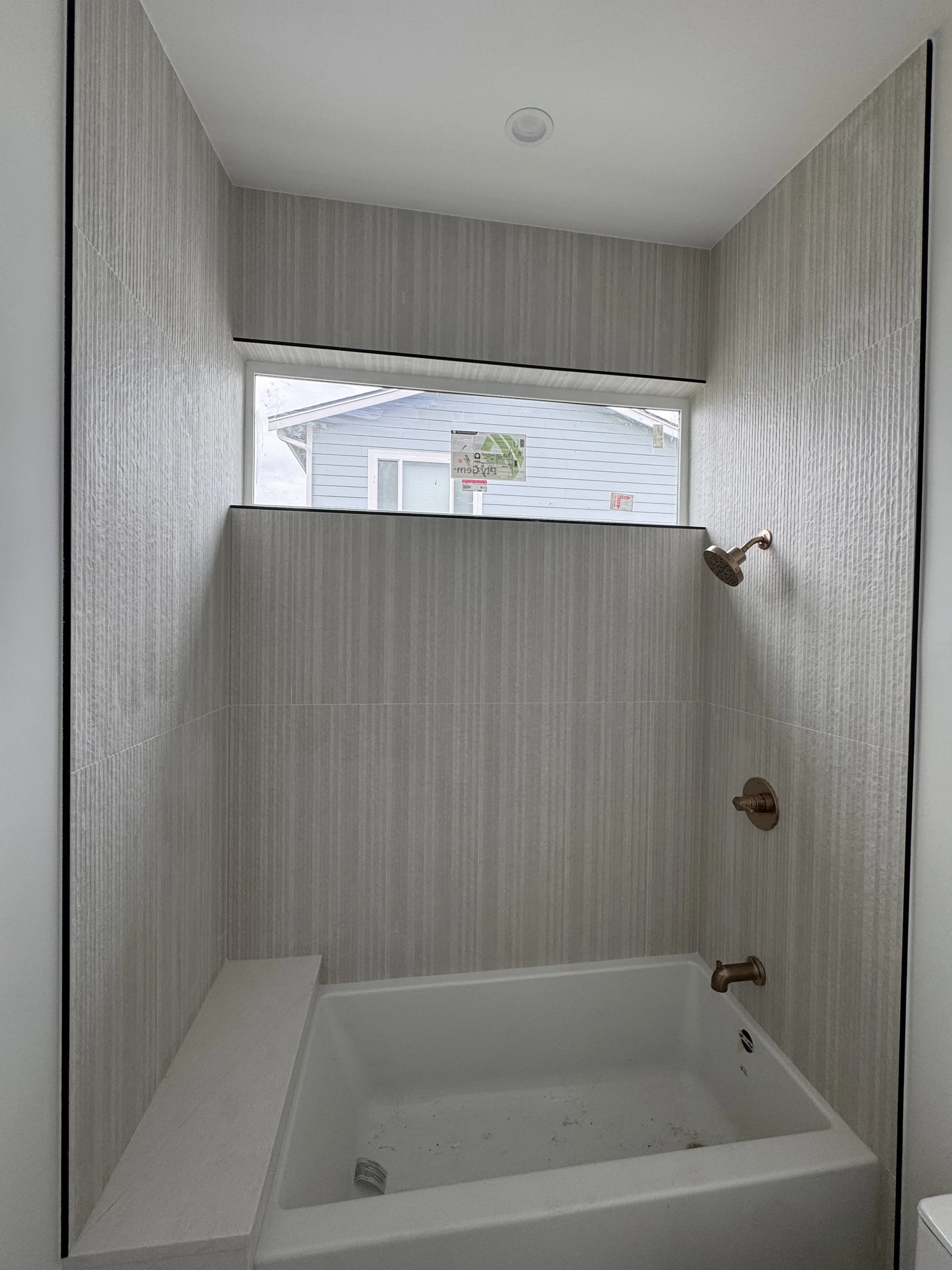 Empty shower with textured beige tile walls, a small built-in shelf, a window with a view of a neighboring house's siding, and bronze fixtures including a showerhead, handle, and drain