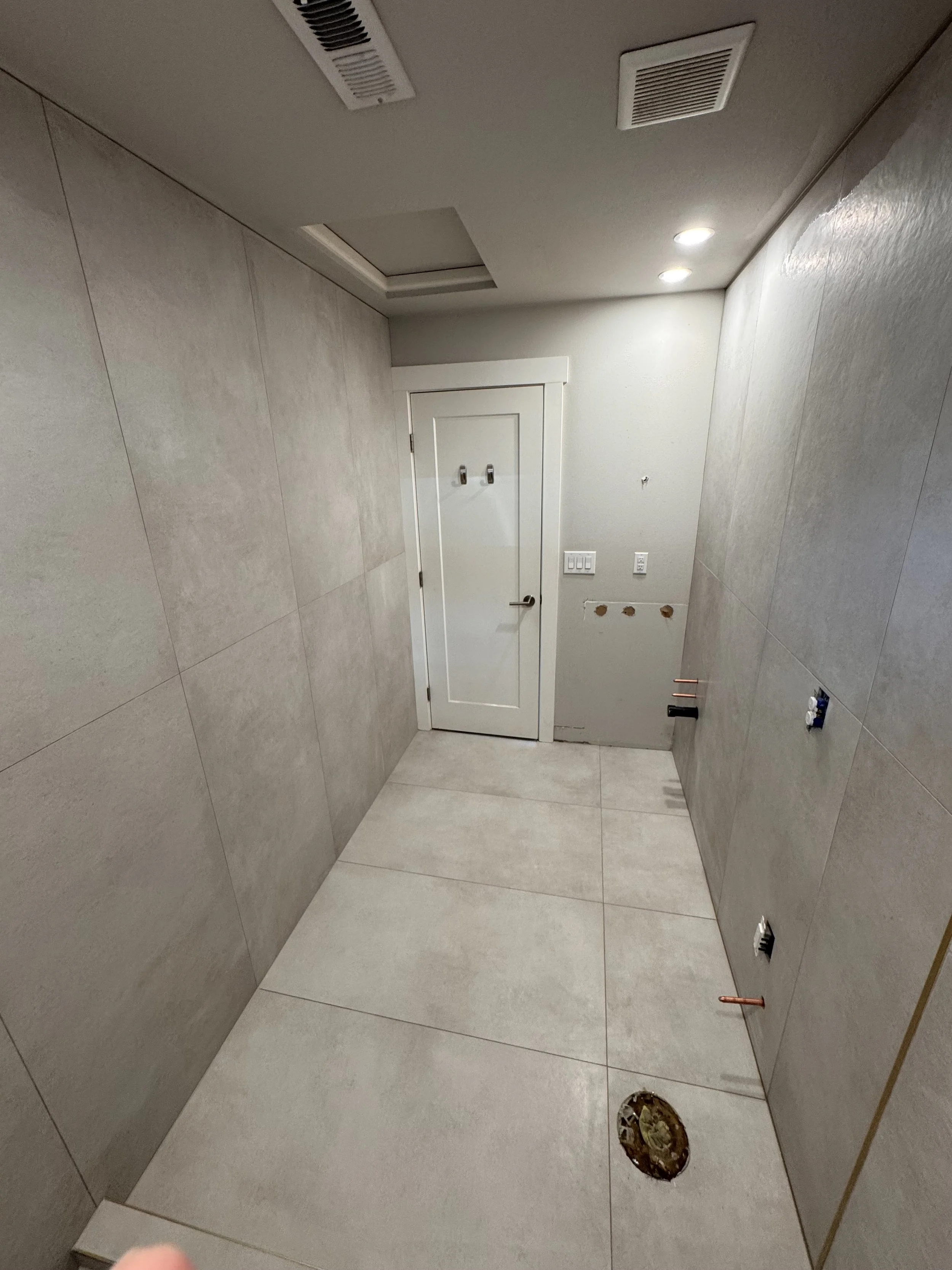 A bathroom or utility room with tiled walls and floor, a closed door with two hooks on it, and plumbing fixtures on the wall. The ceiling has ventilation vents and recessed lighting.