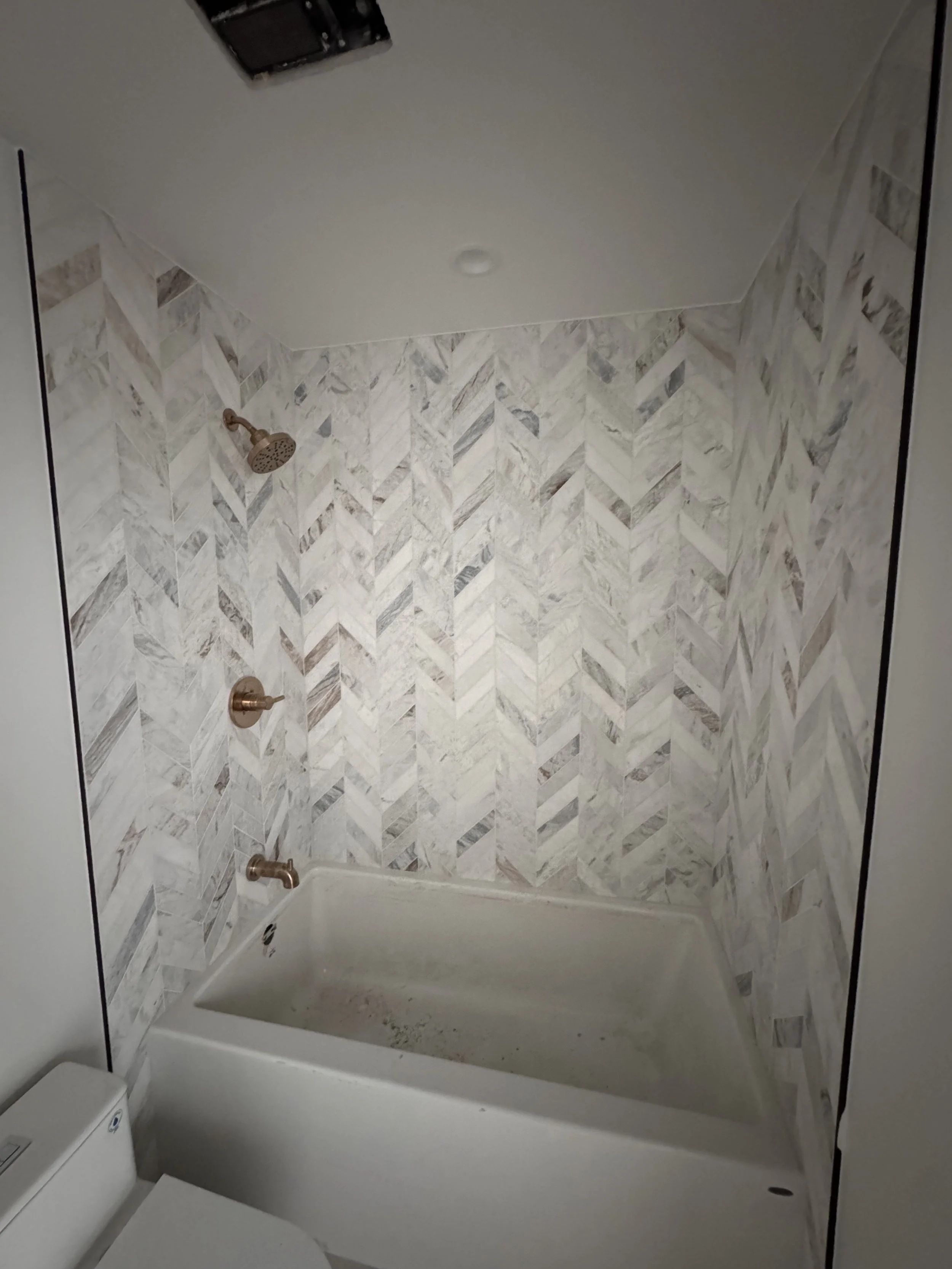 Bathroom shower with marble wall tiles, a showerhead, a control handle, and a small white toilet in the corner.