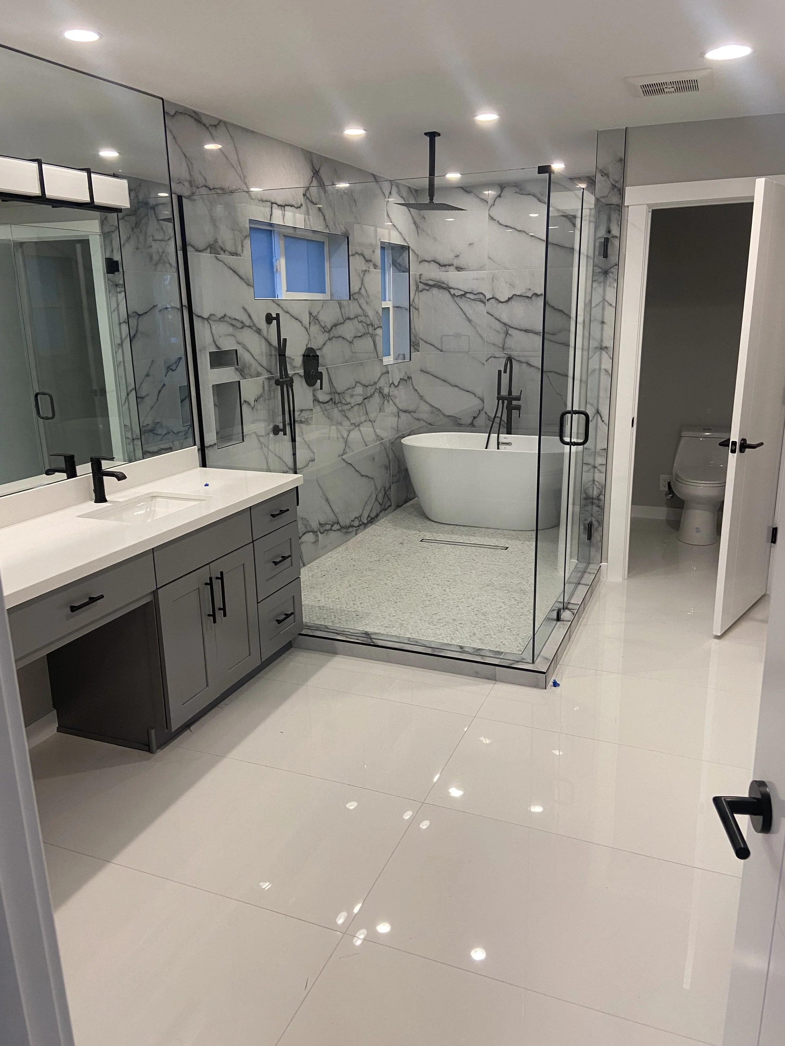 Modern bathroom with large white tiled floor, marble walls, glass-enclosed shower, freestanding bathtub, and a vanity with a white countertop and black fixtures.