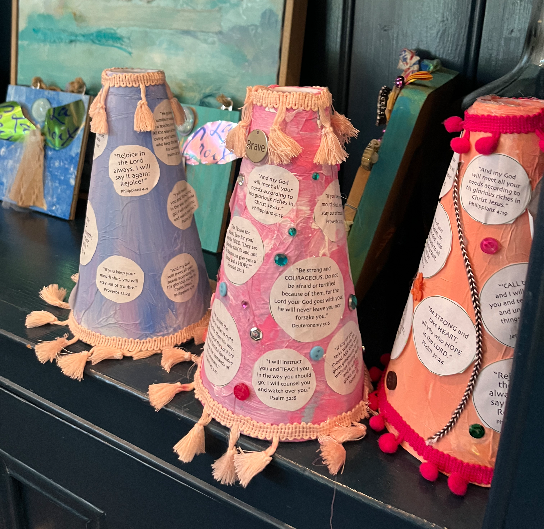 Colorful handcrafted paper lanterns with religious and inspirational messages in paper cutouts, decorated with tassels, pom-poms, and rhinestones, displayed on a dark surface.