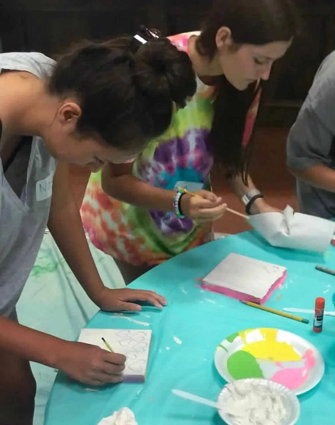 People making colorful tie-dye shirts at a table covered with a blue plastic tablecloth, with bowls of dye, a glue stick, and paper towels.