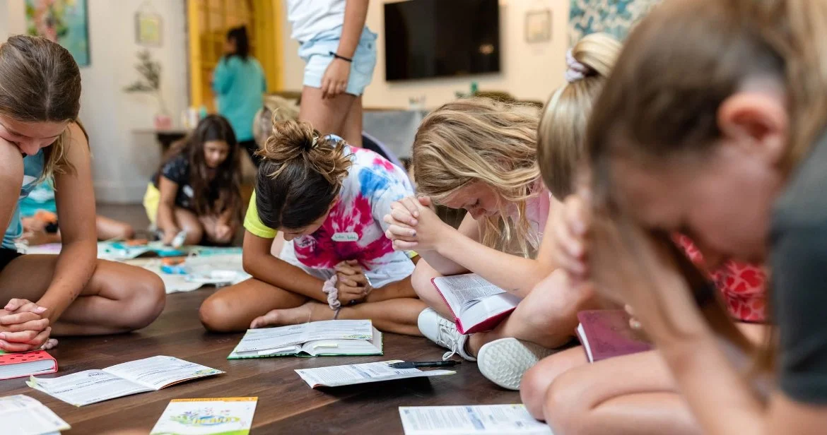 Group of children kneeling on a wooden floor, praying with books open in front of them.