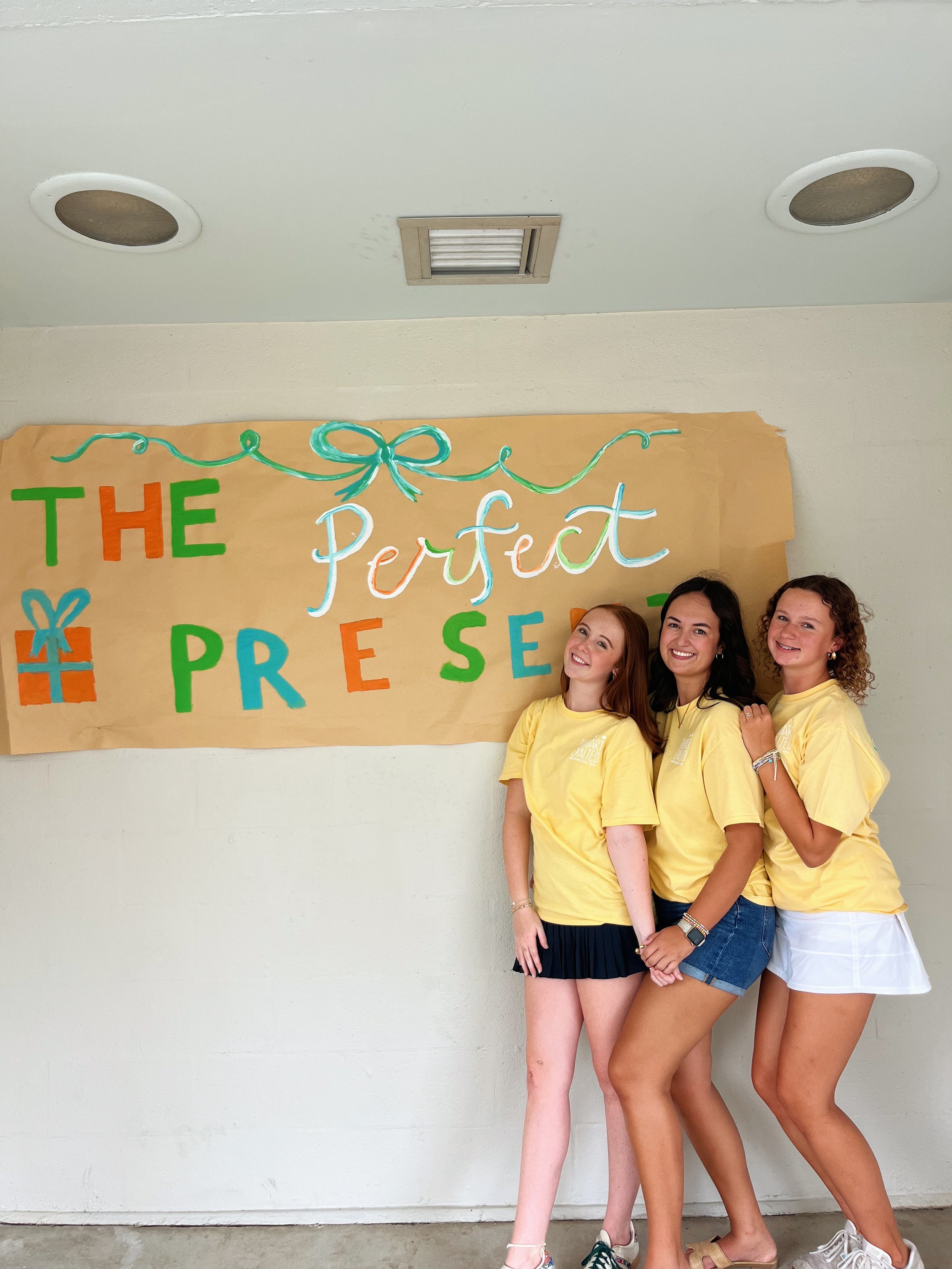 Three women standing in front of a colorful poster on a beige wall, smiling for a group photo. The women are dressed casually in yellow T-shirts and shorts, with one wearing a black skirt and another in white shorts.