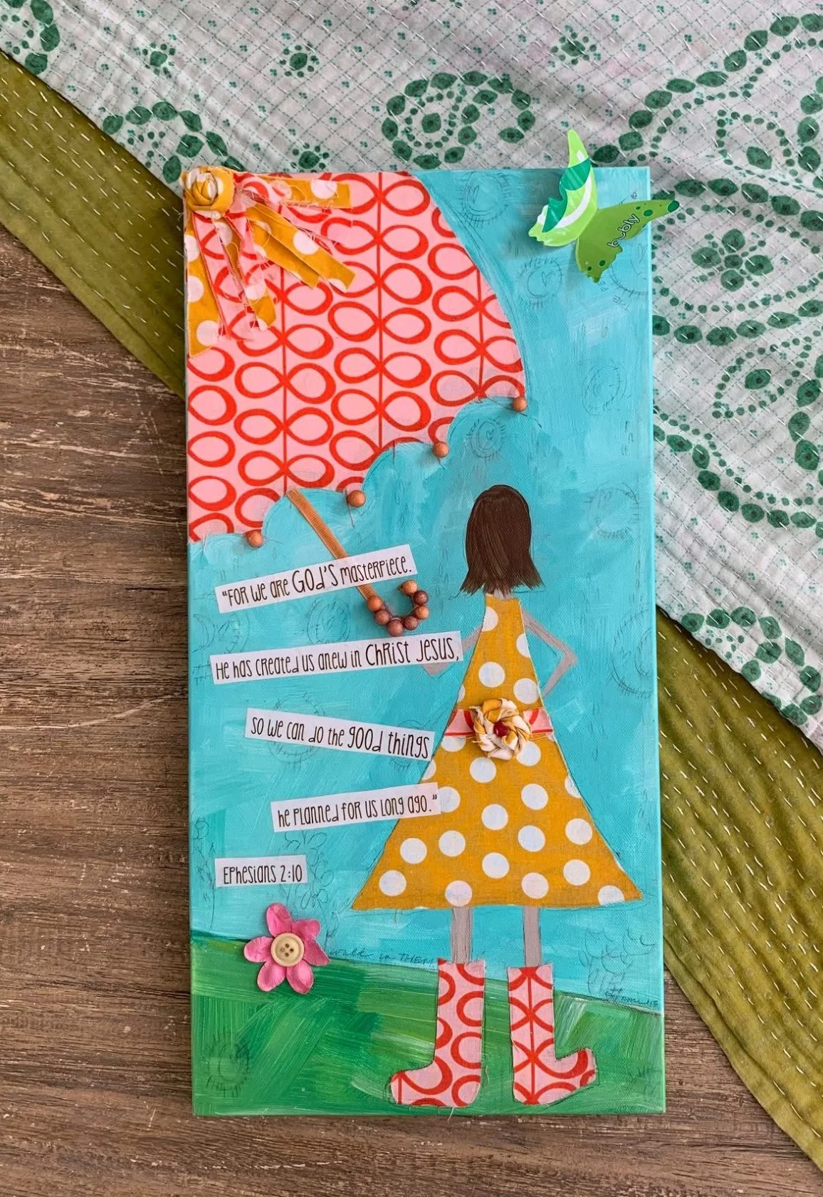 Handmade Christian-themed artwork on paper featuring a girl with brown hair in a yellow polka dot dress and red and white boots, standing on green grass. The background includes a blue sky, a large red and pink patterned umbrella, and decorative elem