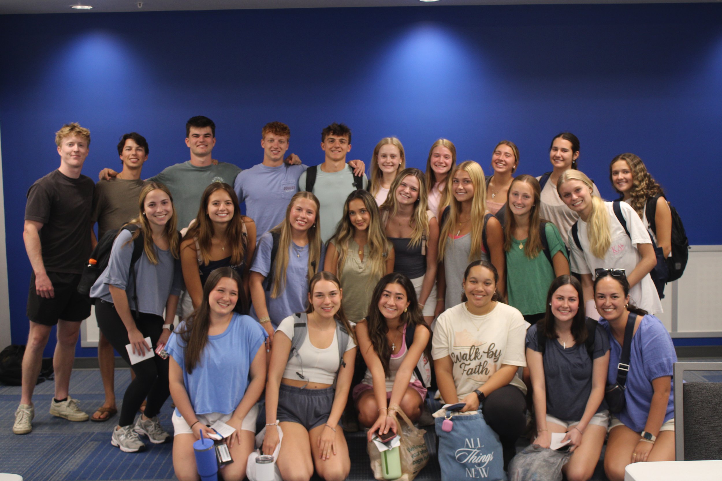 Group of young people, mostly females, smiling and posing together in front of a blue background, some holding notebooks and water bottles.