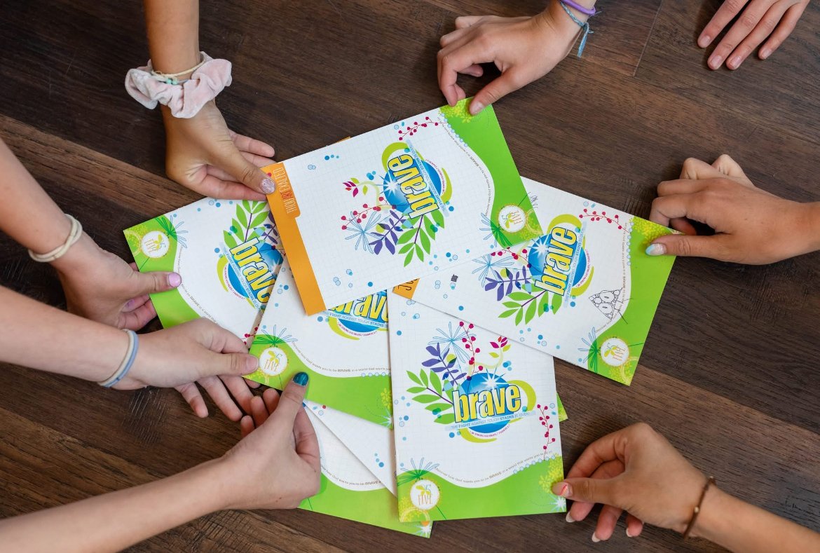 Multiple hands holding and spreading out colorful sheets of paper on a wooden table, with the sheets displaying the word "brave" and decorative patterns.