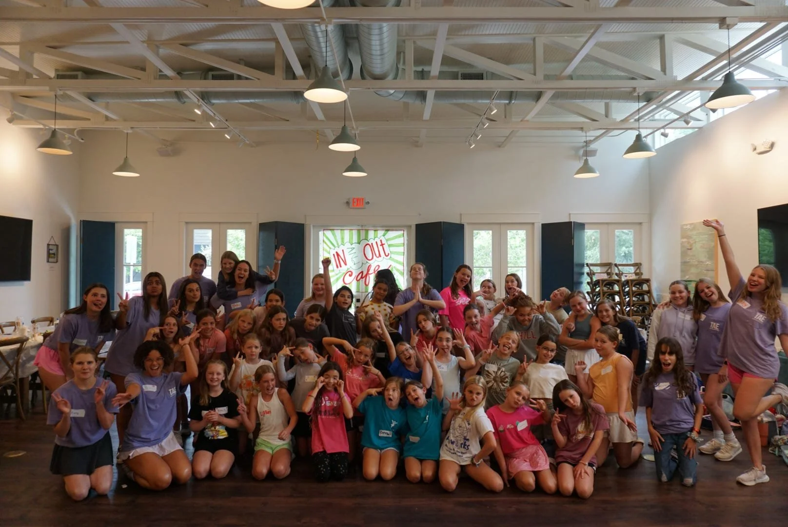 A large group of children and teenagers gathered indoors for a group photo, smiling and making various gestures. The setting appears to be a community or activity center with a sign in the background reading 'In and Out Cafe.'