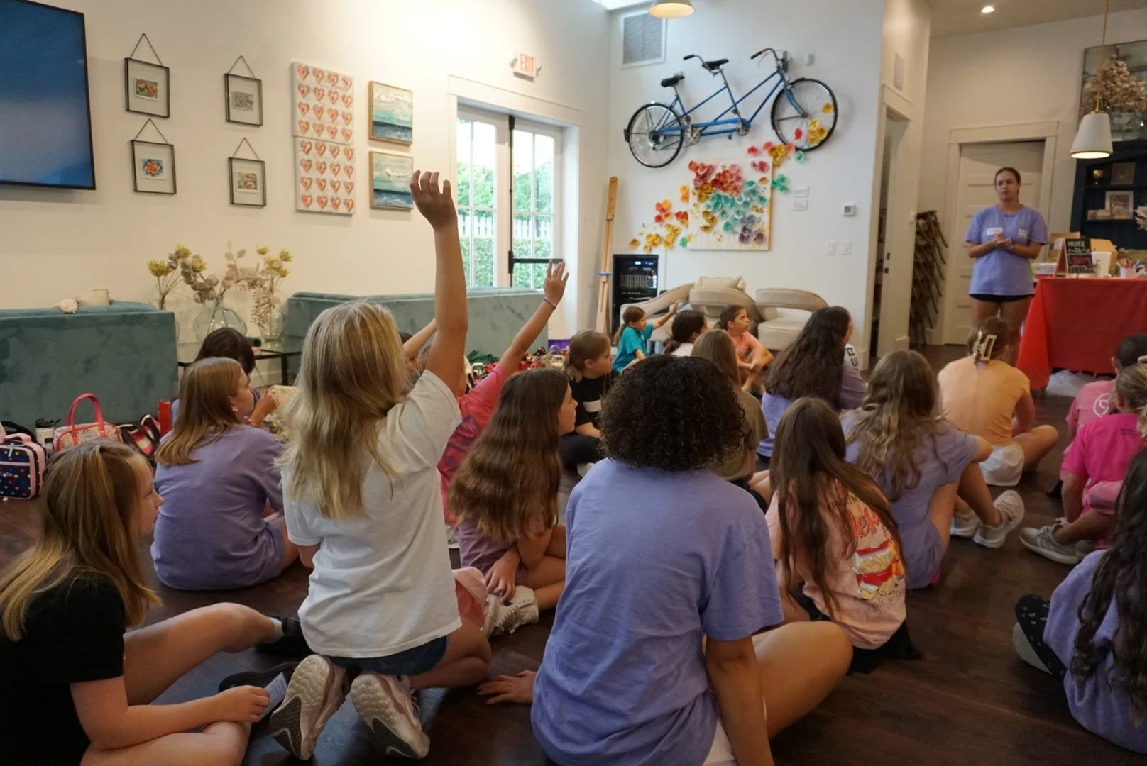 Children sitting on the floor in a room listening to a woman speak. Several children have their hands raised. The room has artwork on the walls, a bicycle hanging on the wall, and a door with a window letting in natural light.