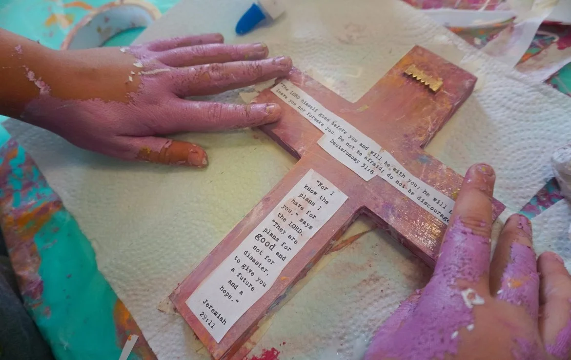 Hands with paint on fingers working on a decorated pink cross with biblical text and a tag resembling a prayer or message.