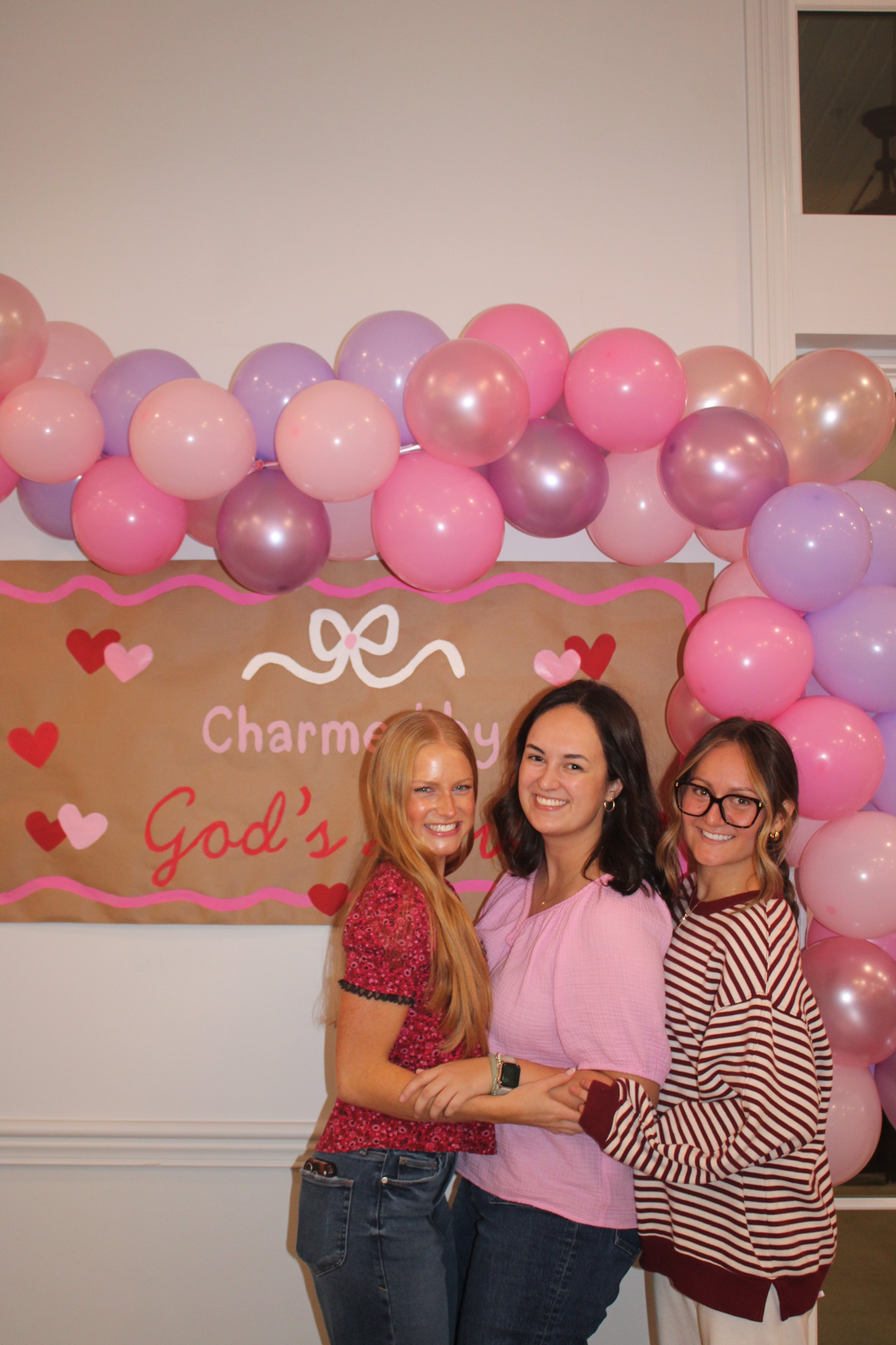 Three women smiling and standing together in front of a pink and purple balloon arch and a Valentine's Day themed poster that says 'Charmed By God' with red and pink hearts.