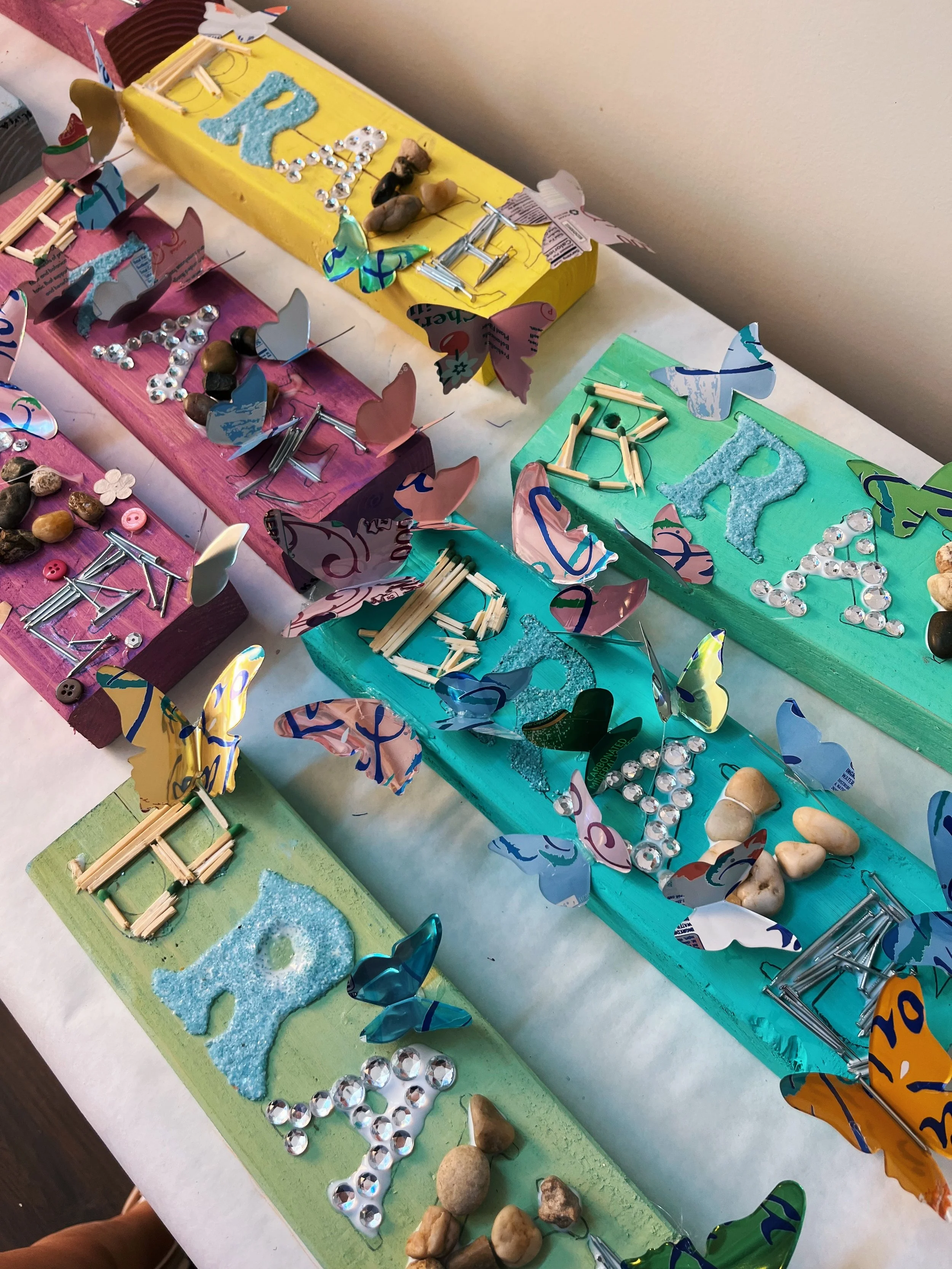 Colorful tables with various craft materials, including painted rocks, butterfly cutouts, rhinestones, buttons, sticks, and glitter, arranged for a crafting activity.