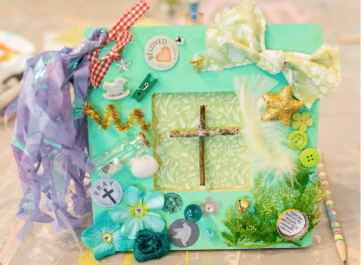 Decorated picture frame with religious and nature-themed embellishments, includes a cross made from sticks, flowers, buttons, bows, sequins, and stickers with messages like 'Loved' and 'Pray'.