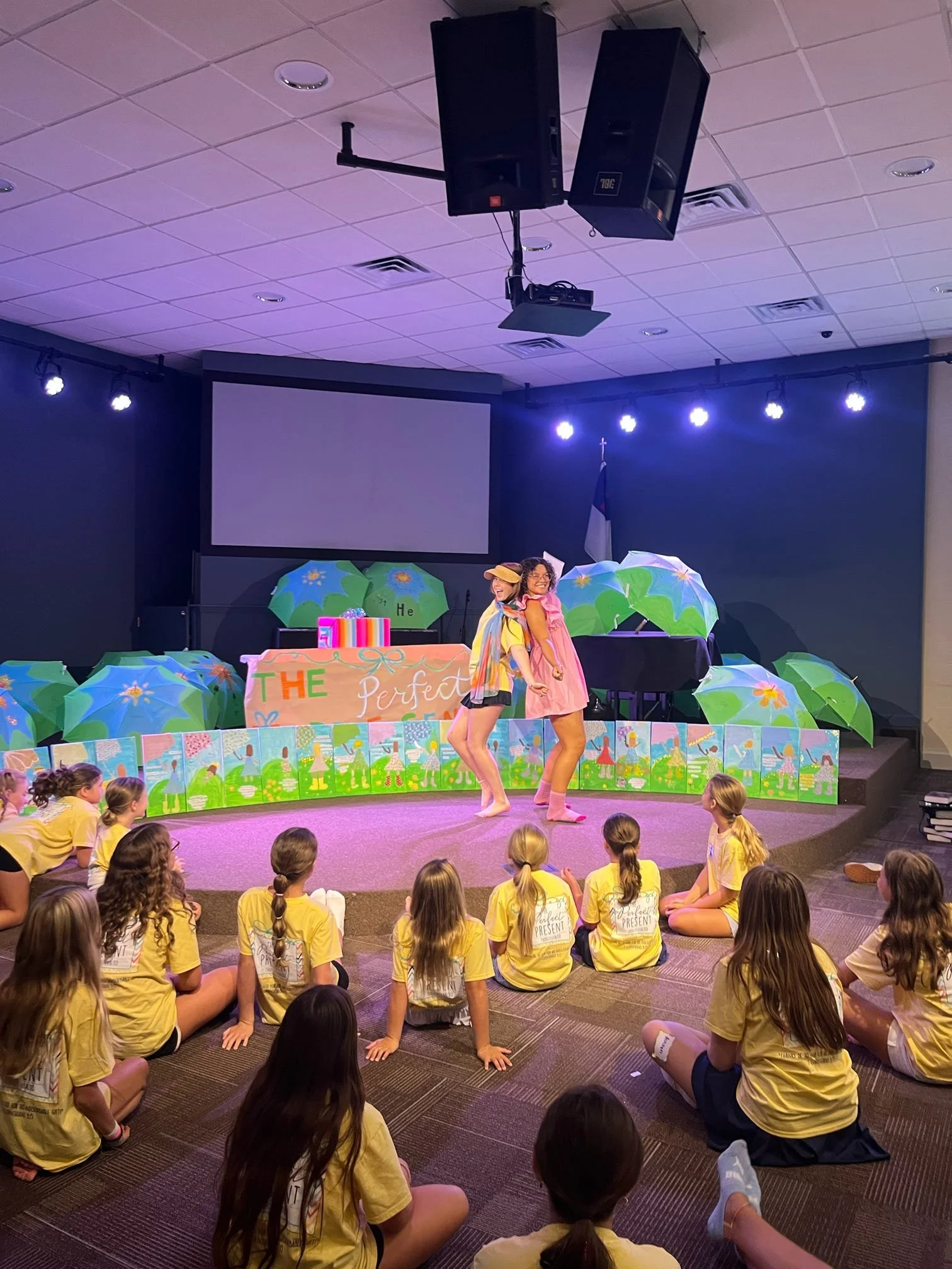 Children sitting on the floor and on stage watching a performance with two dancers in yellow and pink clothes. The stage is decorated with umbrellas, and there is a large blank screen on the stage. The children are wearing matching yellow t-shirts.