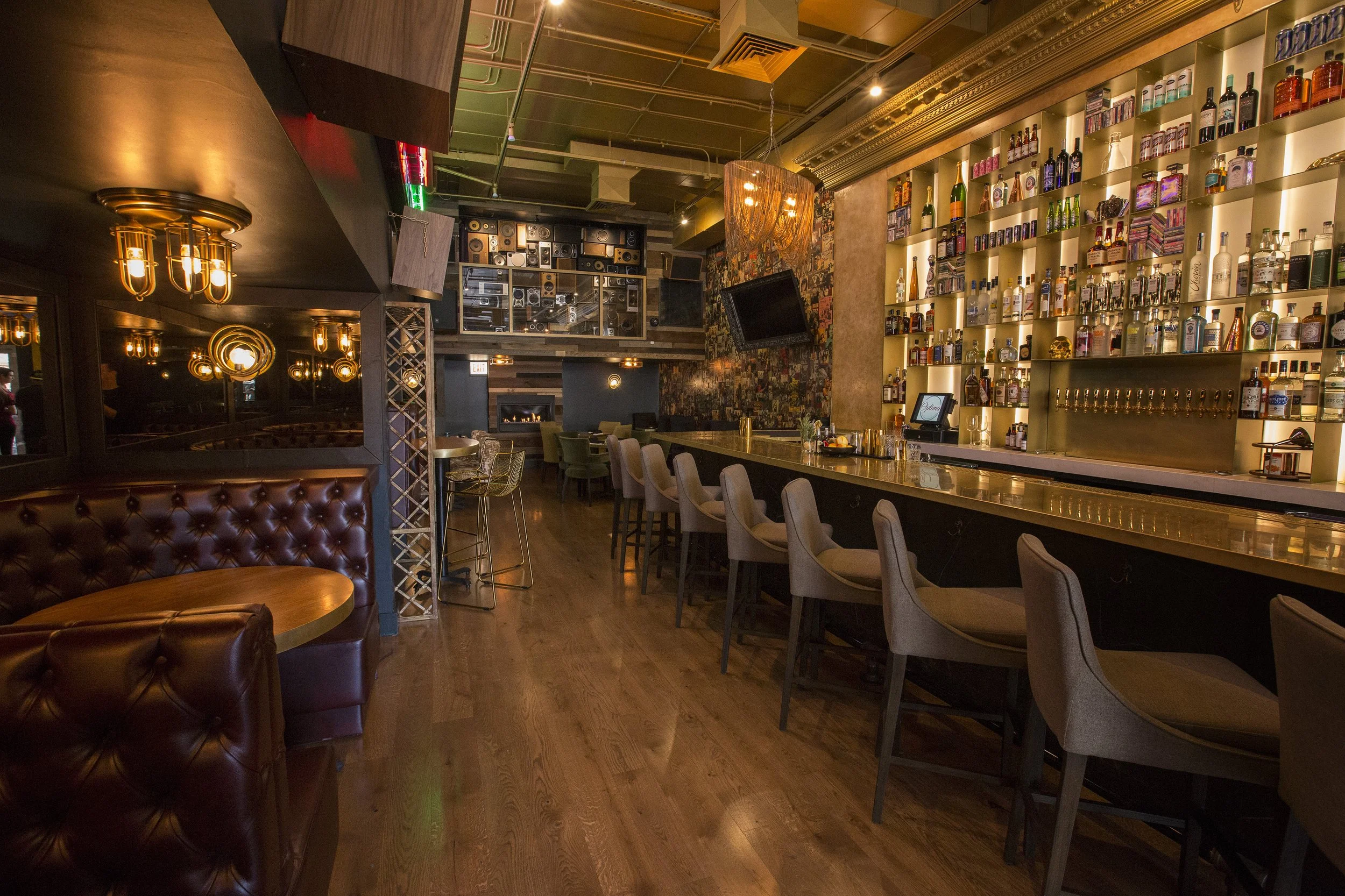 Interior of a modern bar or restaurant with warm lighting, a long bar counter with bar stools, shelves stocked with liquor bottles, a wall-mounted TV, and a decorative chandelier.