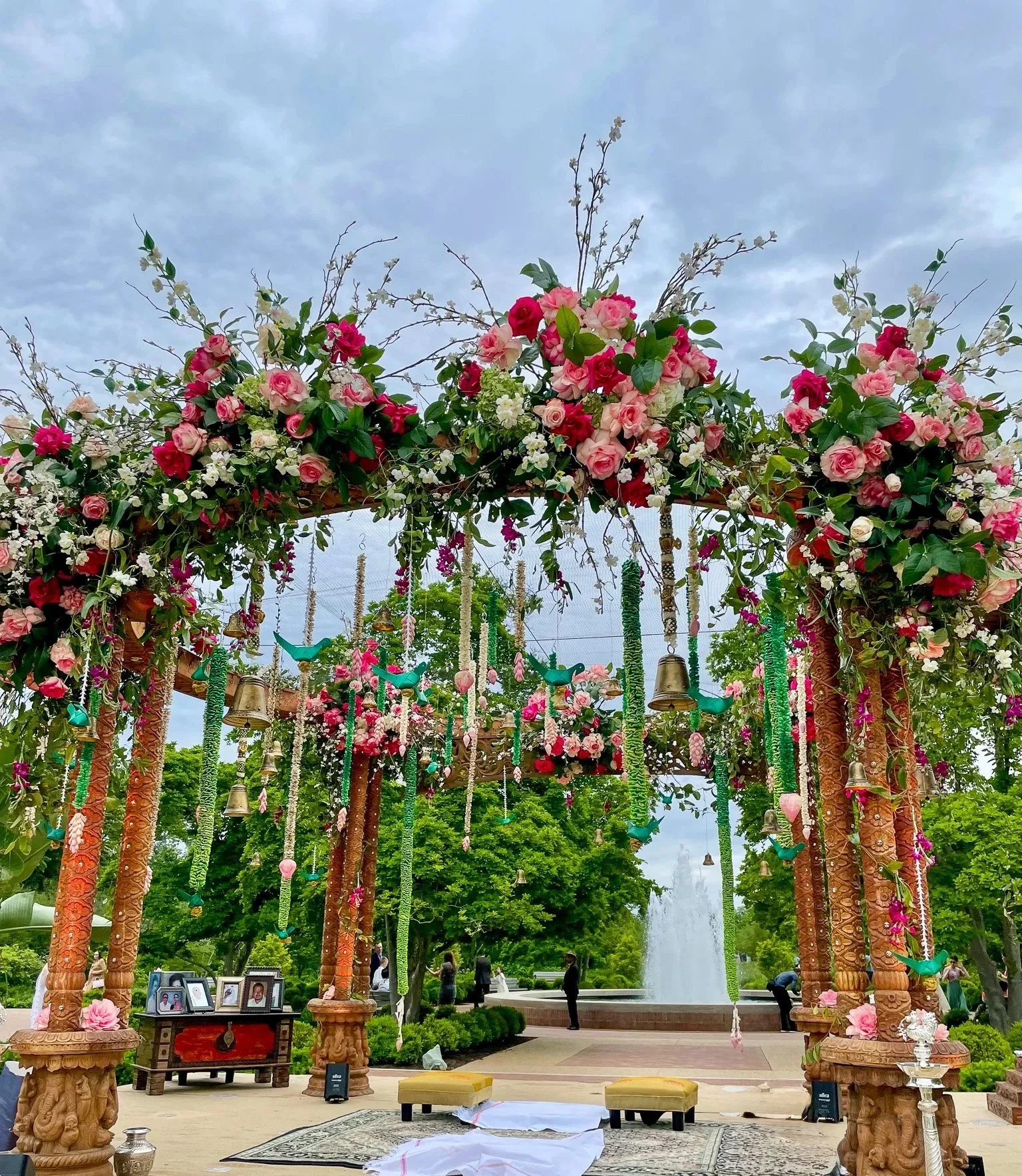 A decorative outdoor structure with carved wooden pillars decorated with flowers and hanging bells, set in a park with trees and water fountain in the background.