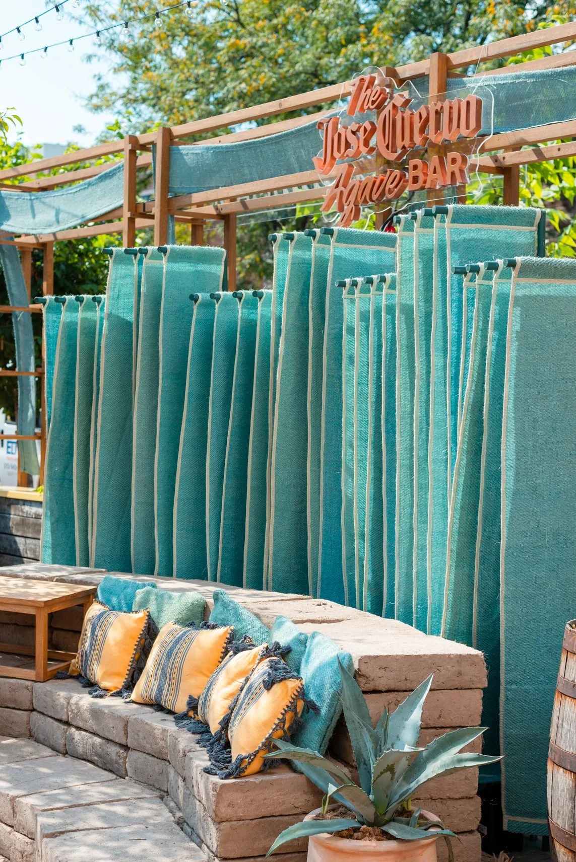 Outdoor display of turquoise striped textiles hanging on a wooden frame, with a sign that reads 'The Jose Ochoa Have Bar,' accented with string lights, yellow pillows, and potted plants.