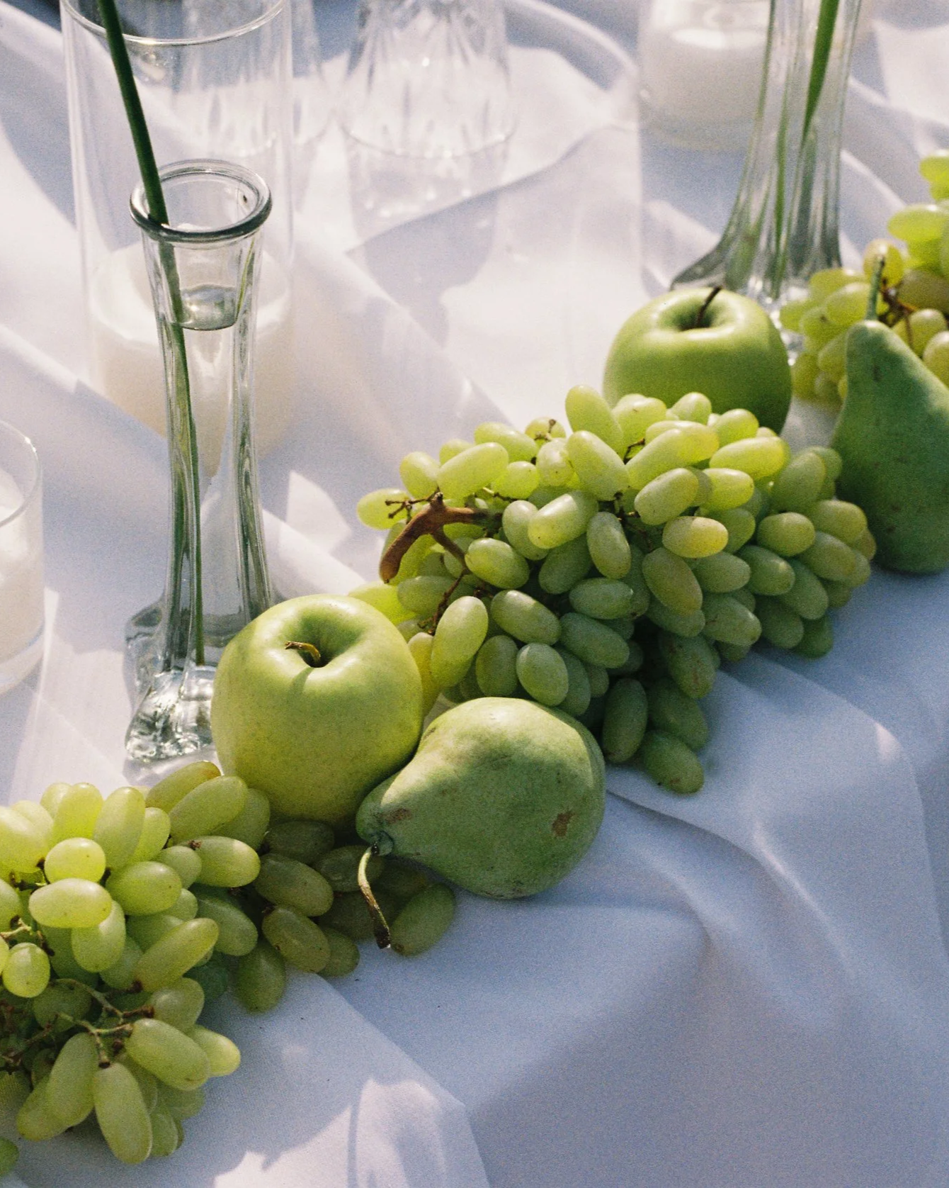 A table decorated with green apples, green pears, bunches of green grapes, clear glass vases, and empty glasses on a white tablecloth.