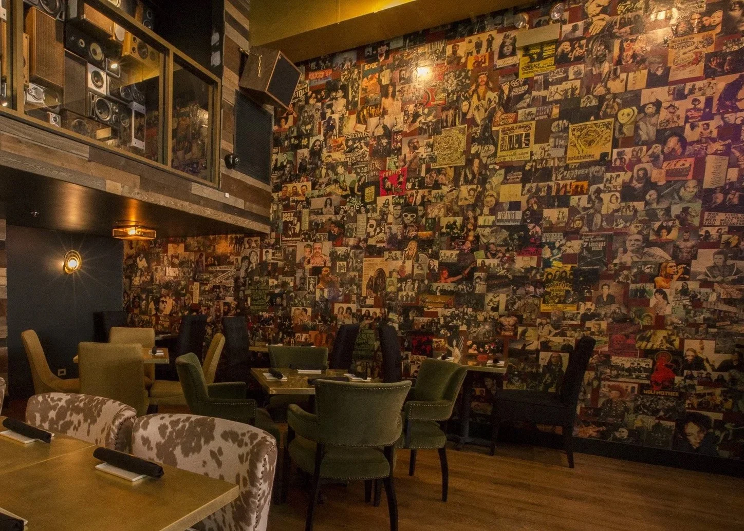 A restaurant or bar with a wall covered in numerous vintage concert and album covers. There are tables with chairs and napkins in the foreground, and a sound system or speaker on a balcony above.